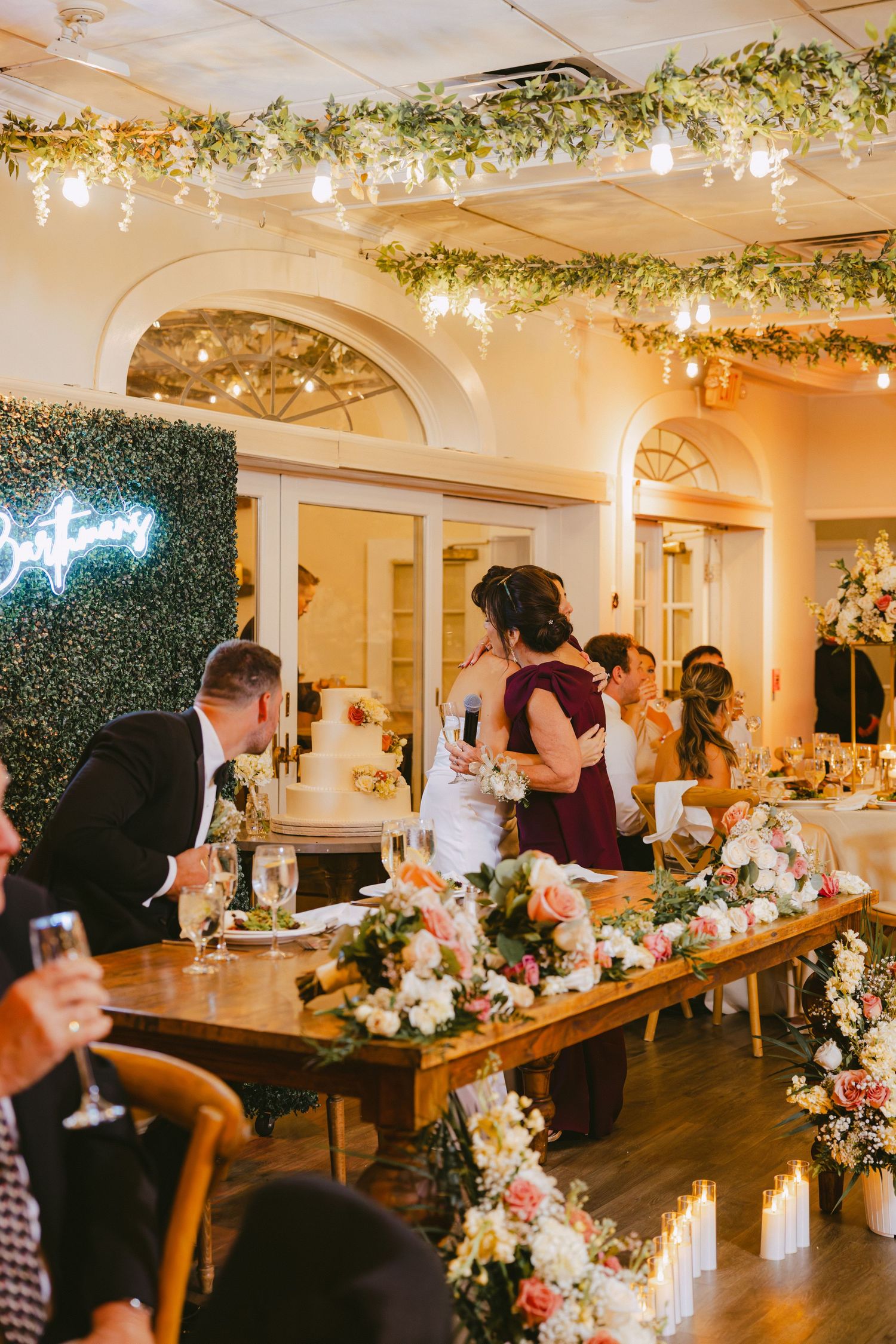 Bride embraces a guest in burgundy at a romantic wedding reception with greenery decor.