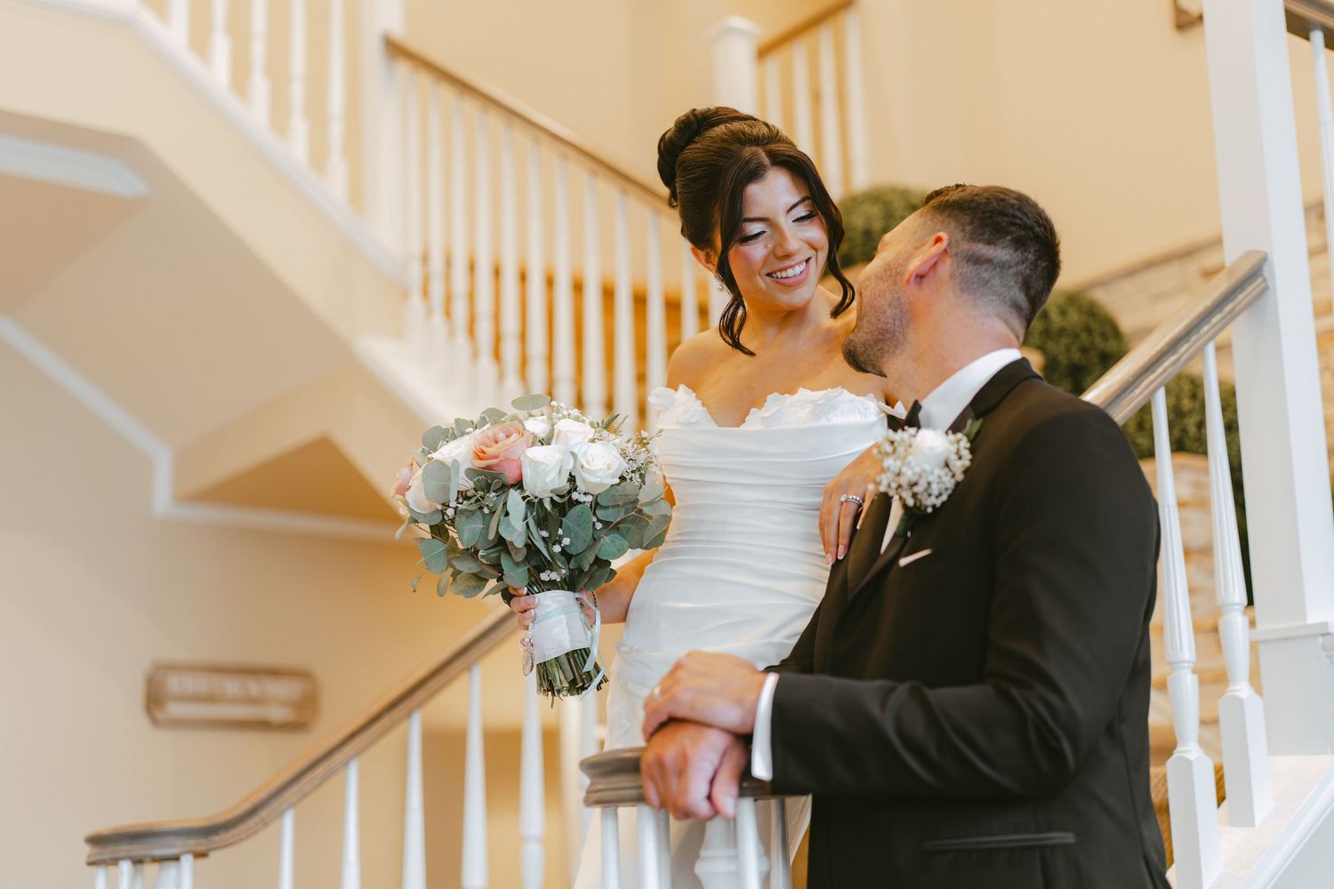 Bride laughing with groom on a grand staircase while holding a soft pink and white bouquet.