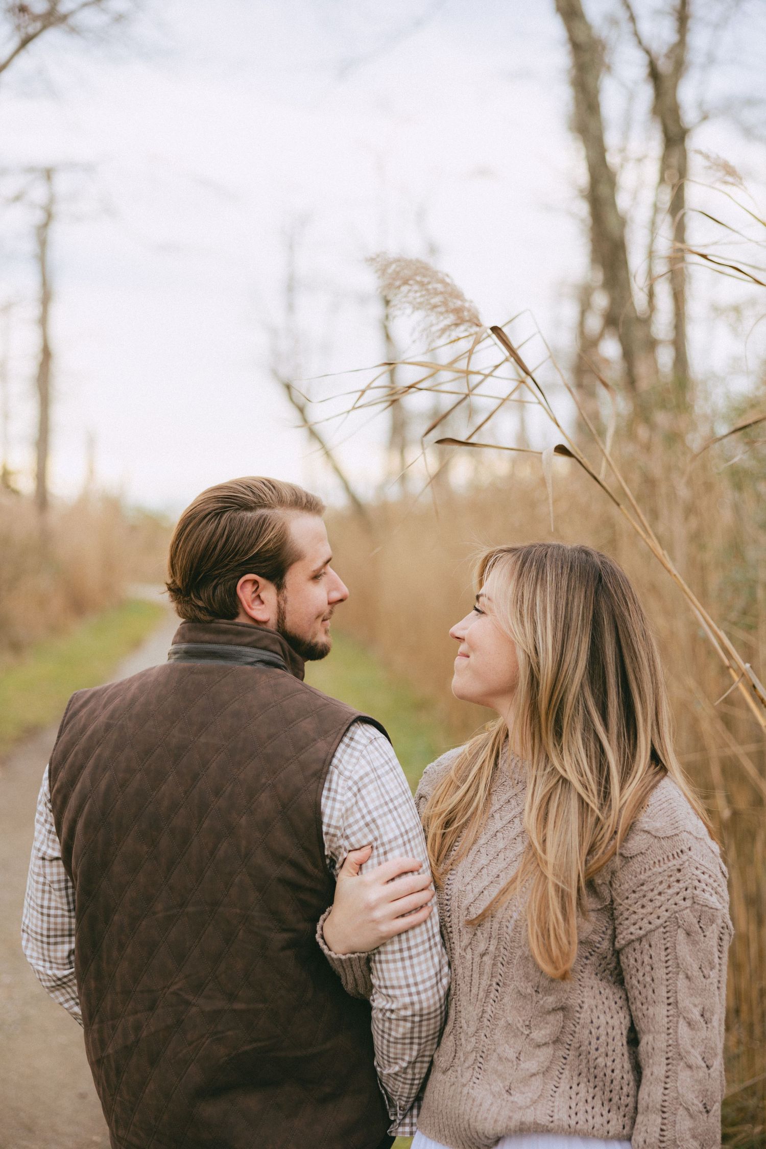 Engaged couple walking on a path surrounded by autumn trees, woman smiling up at her partner.