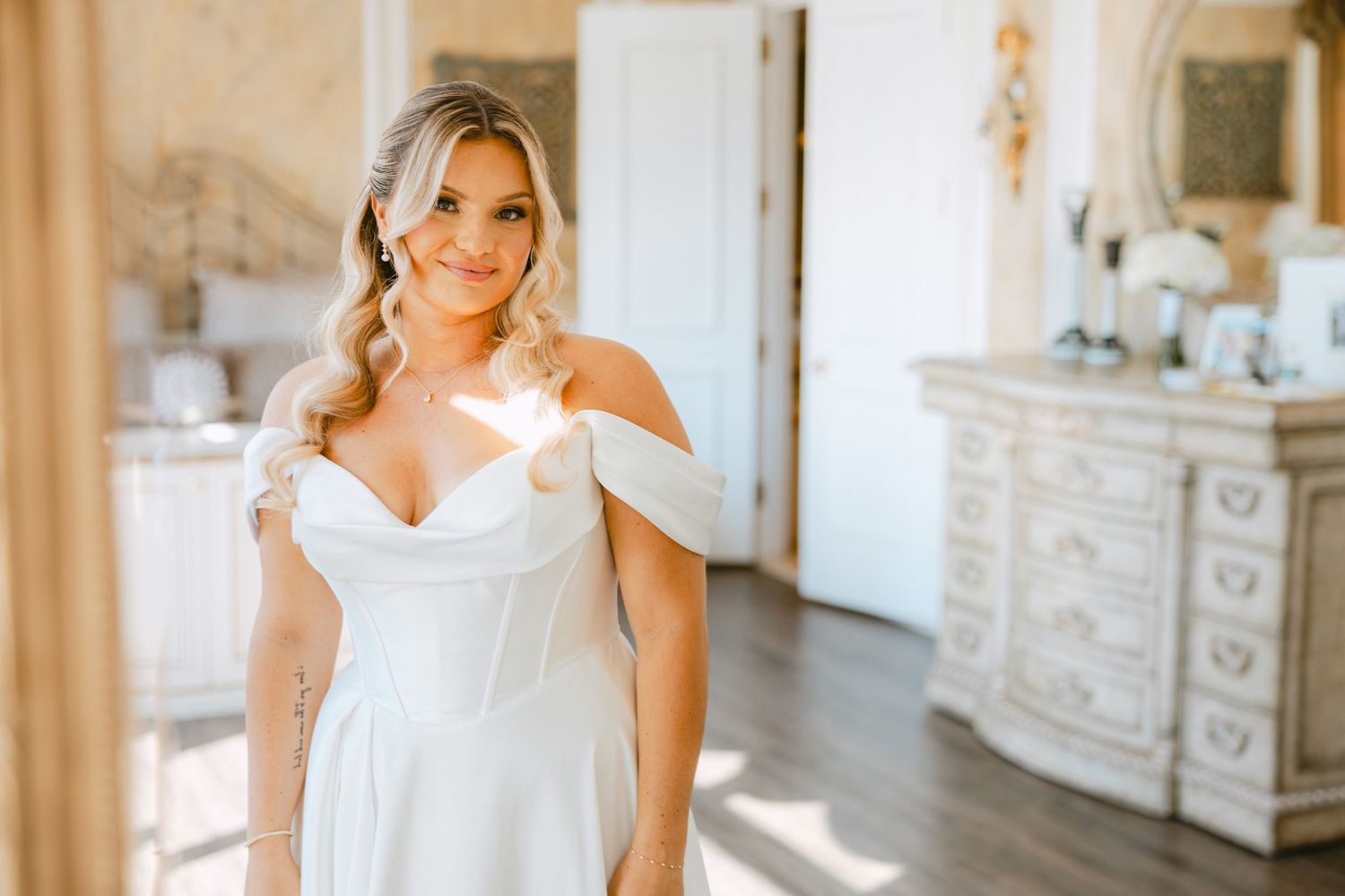 Beautiful bride in off-shoulder white wedding gown poses in elegant sunlit bridal suite.