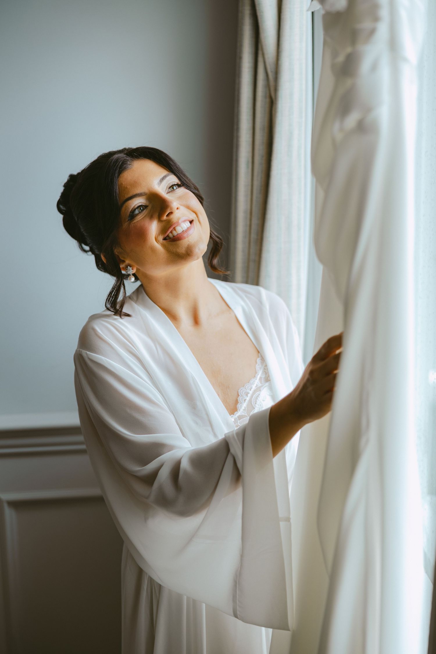 Bride in white robe smiling while touching her wedding dress hanging near a curtained window.