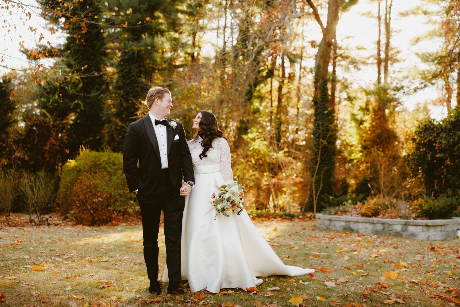 Bride and groom share tender moment in golden autumn garden setting with colorful foliage.