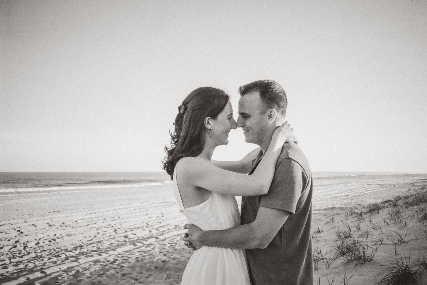 Black and white photo of a couple embracing and smiling on a sandy beach with dunes behind them.