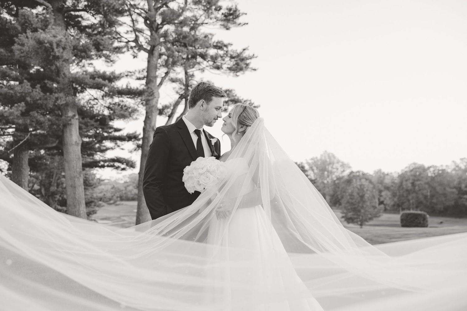 Black and white romantic photo of bride and groom kissing with veil flowing in the wind among tall trees.