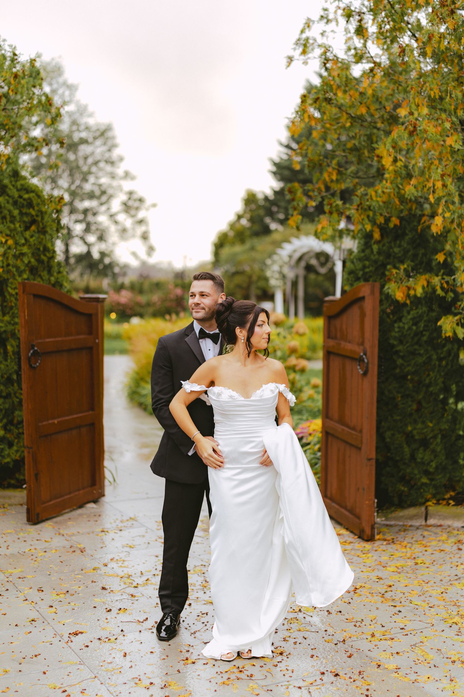 Groom embracing bride from behind as they walk through open wooden garden gates in autumn.
