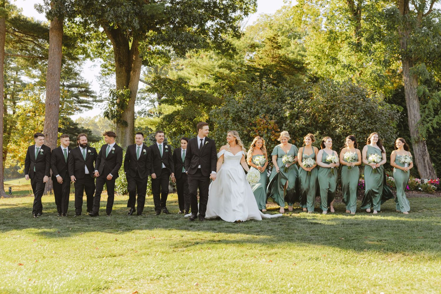 Wedding party poses outdoors with bride, groom, groomsmen in black, and bridesmaids in sage green.