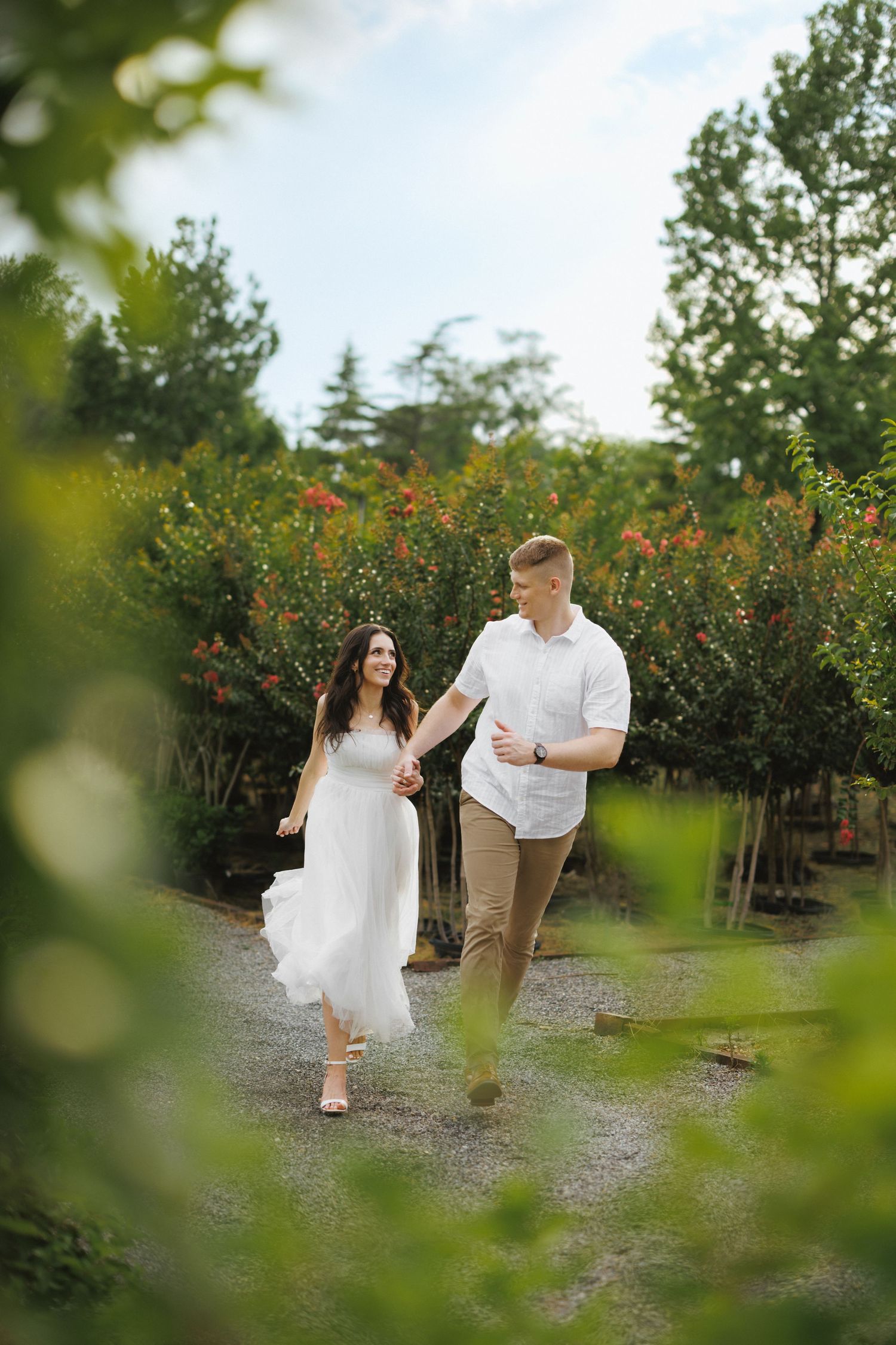 A couple holds hands walking through a lush garden path, woman in white dress, man in white shirt and tan pants.