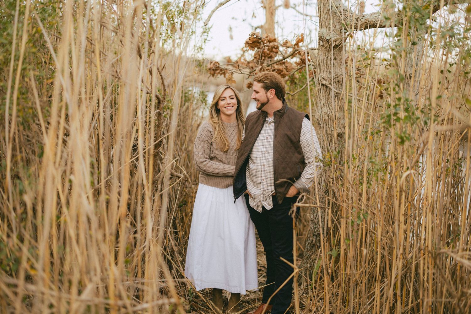 Happy engaged couple standing together among tall golden grasses in an autumn outdoor setting.