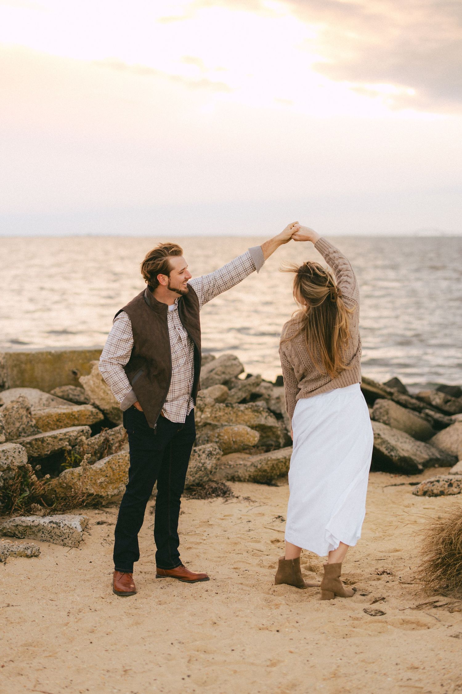 Man spins woman by the hand on sandy beach at sunset with rocky shoreline and ocean in background.