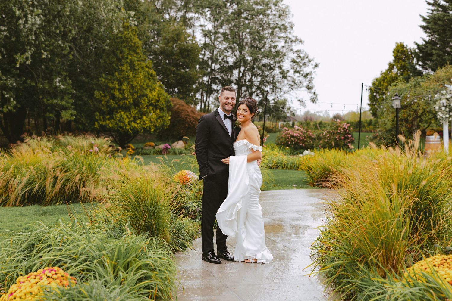 Bride and groom smiling together on a rainy garden pathway surrounded by autumn foliage.