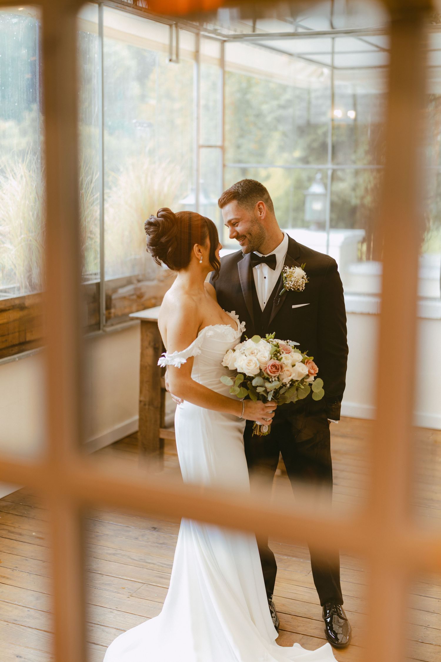 Bride and groom sharing an intimate moment framed by rustic windows in a greenhouse venue.