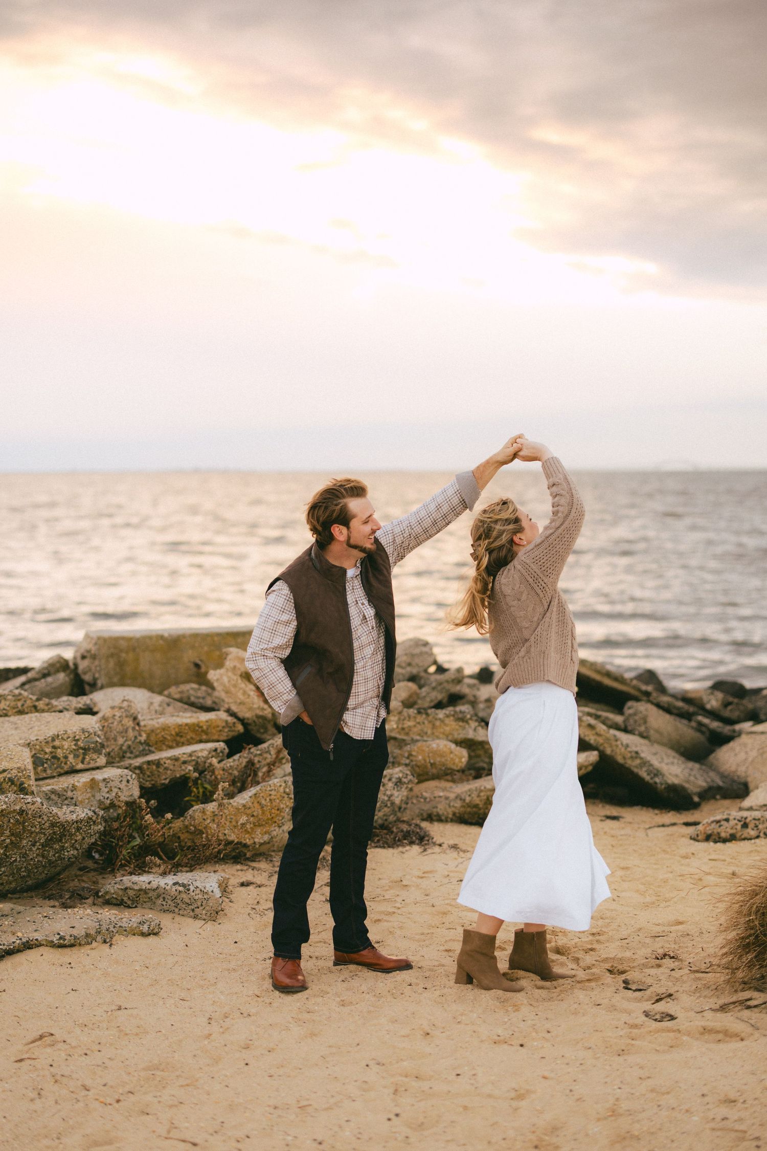 Couple dances on sandy beach at sunset, man spinning woman in white skirt near rocky shoreline and ocean.