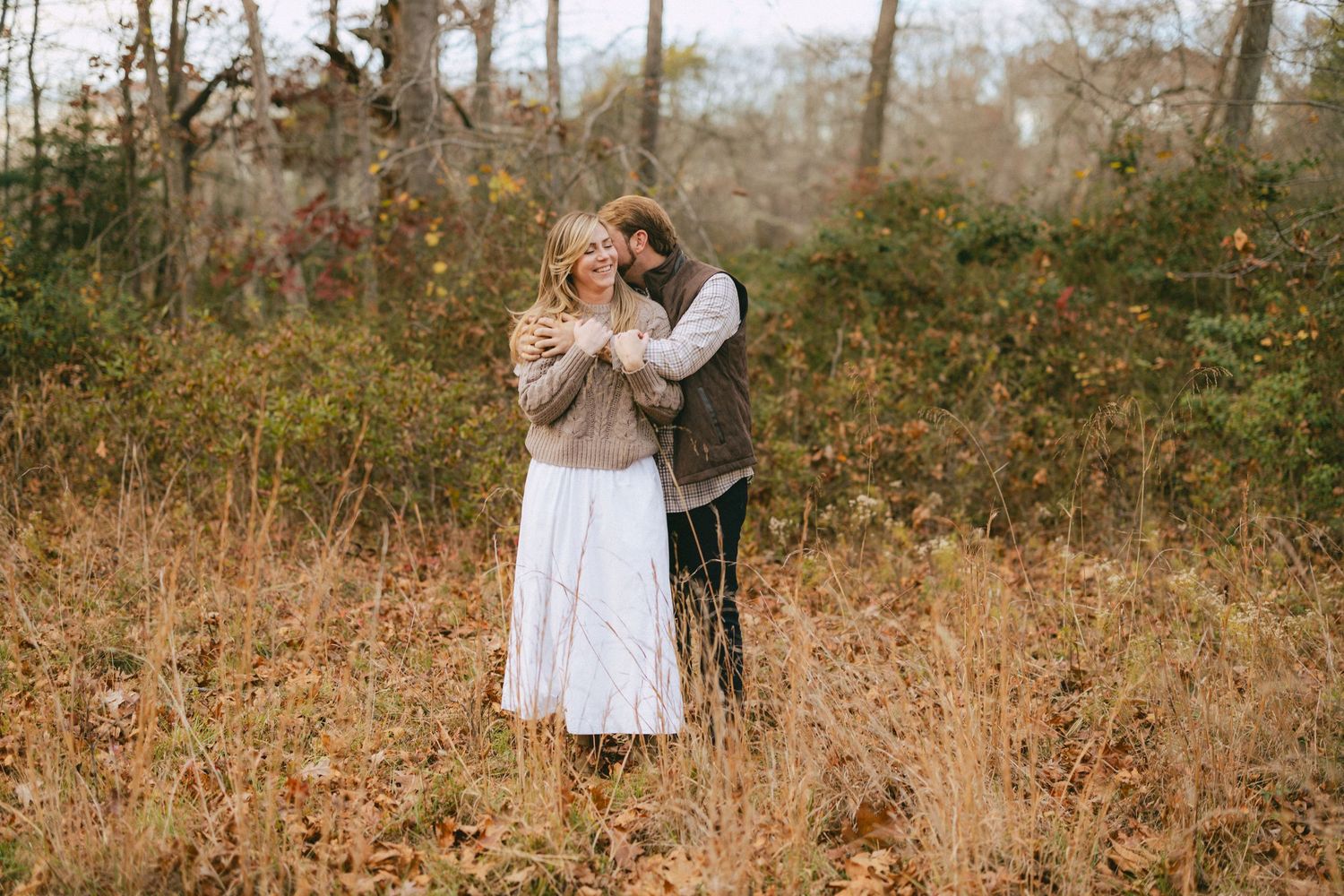 Couple cuddling closely together in a rustic autumn meadow with colorful fall foliage behind them.