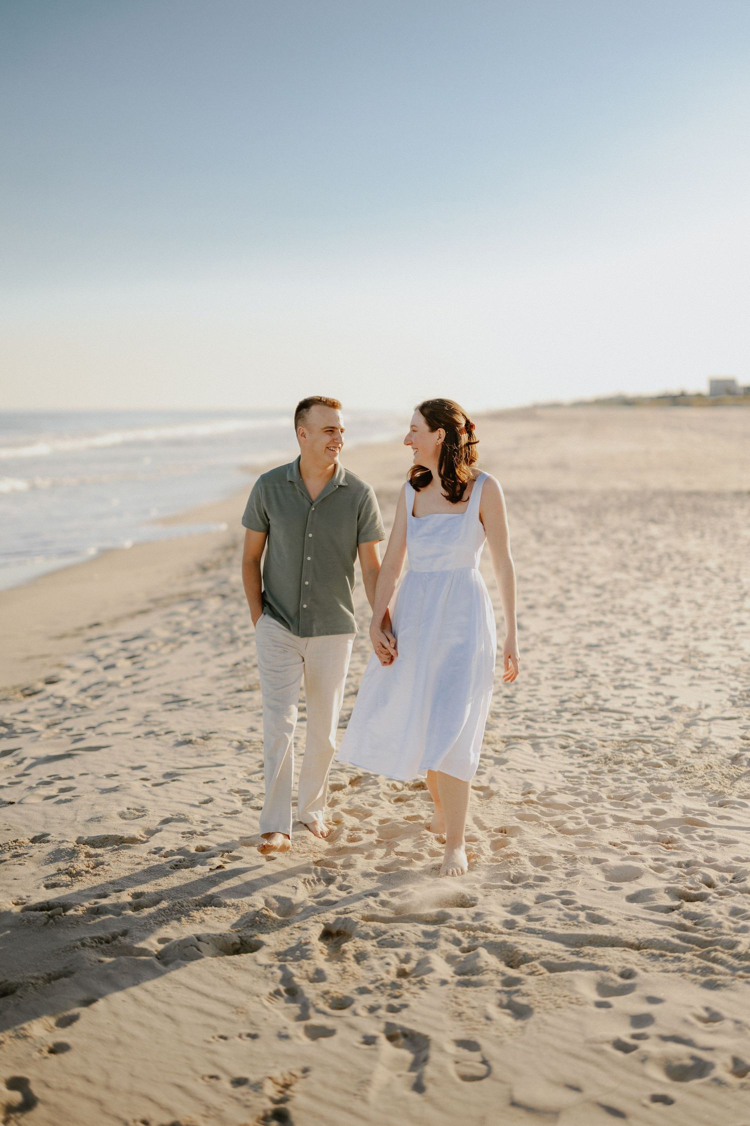 Smiling couple walking hand in hand barefoot along sandy beach shore in warm golden sunlight.