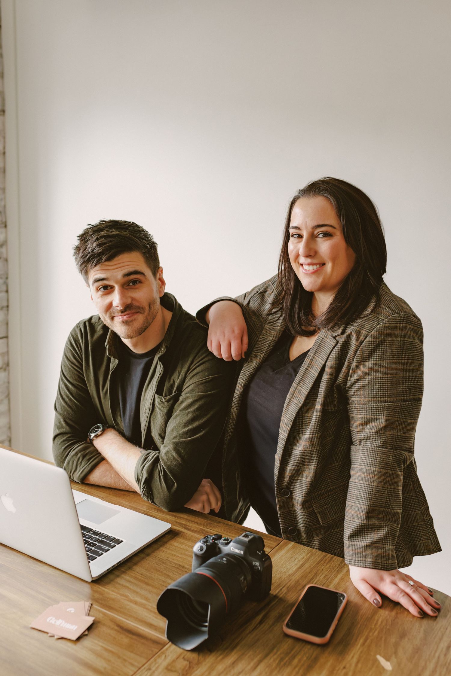Male and female photography duo pose together at desk with camera and laptop in bright studio.