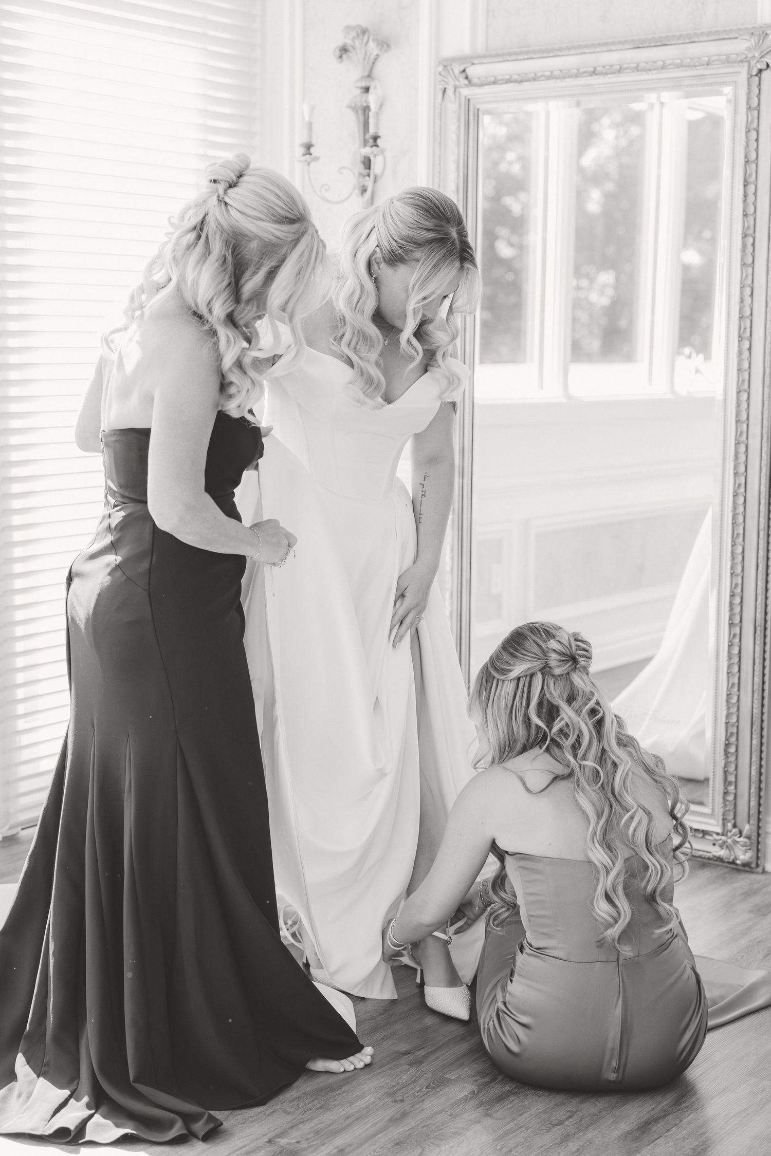 Black and white photo of bridesmaids in dark gowns helping bride into her wedding dress before the ceremony.