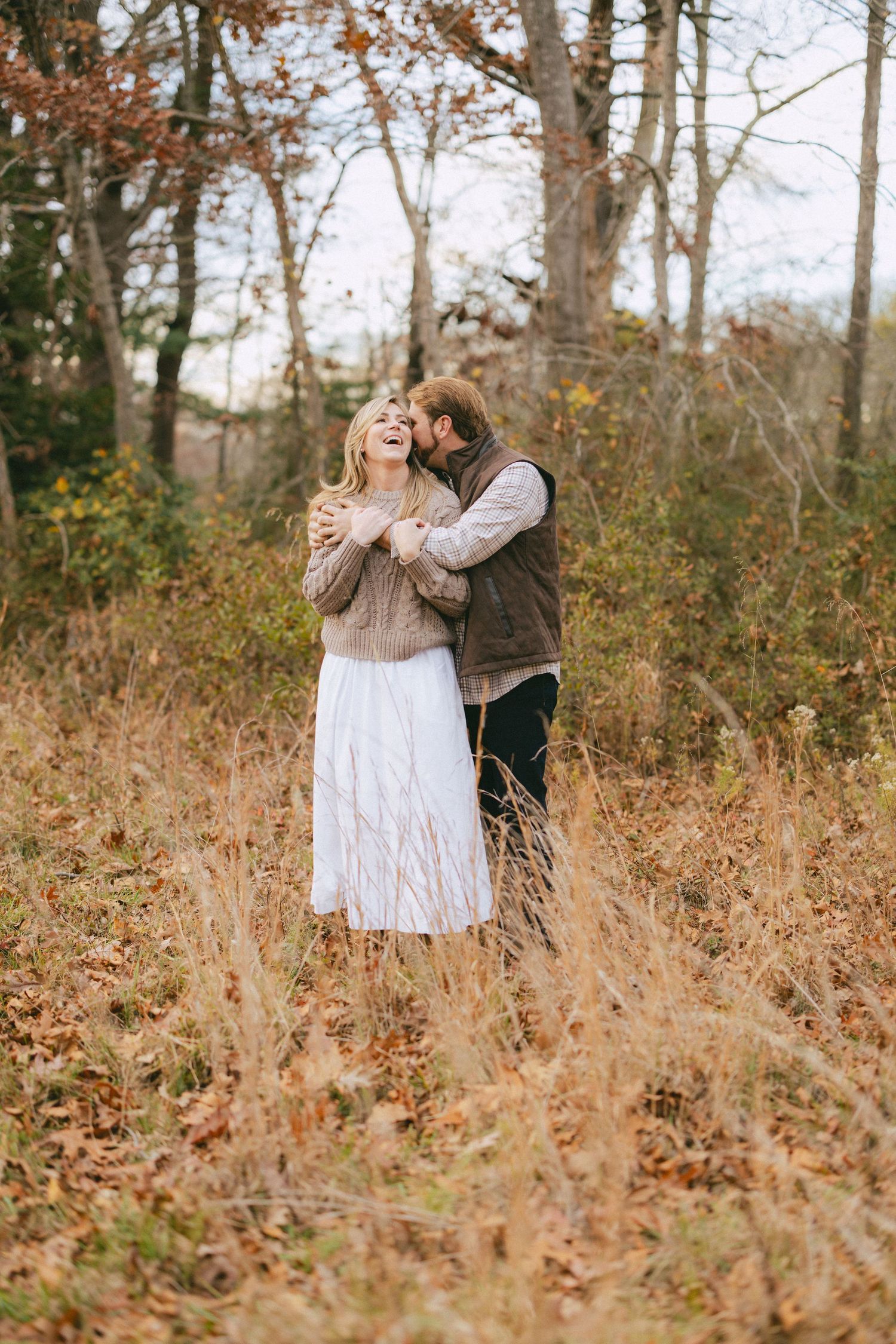 Man embracing laughing woman from behind in a golden autumn field surrounded by fall foliage.