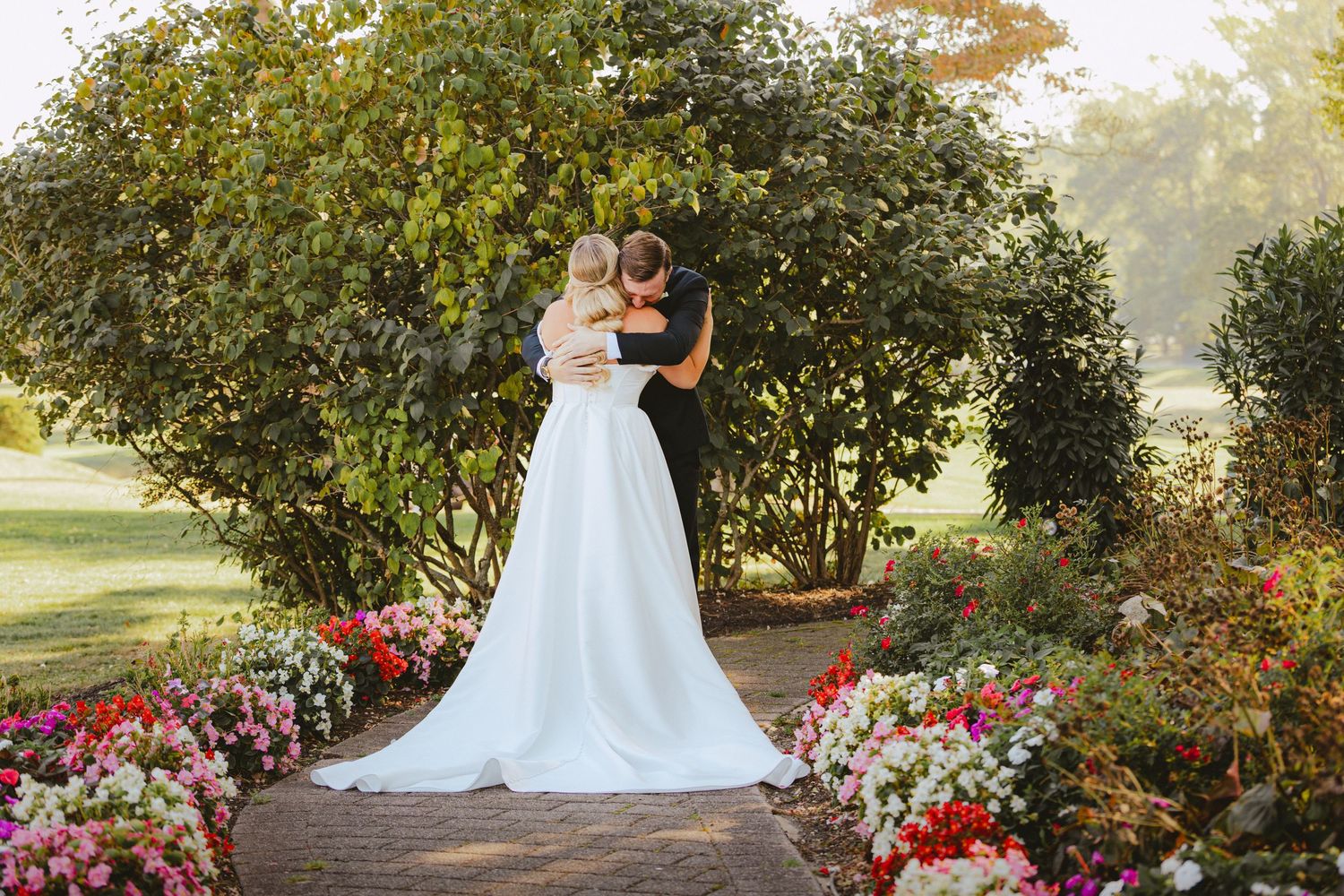 Bride and groom embrace in a romantic garden walkway surrounded by colorful blooming flowers and lush greenery.