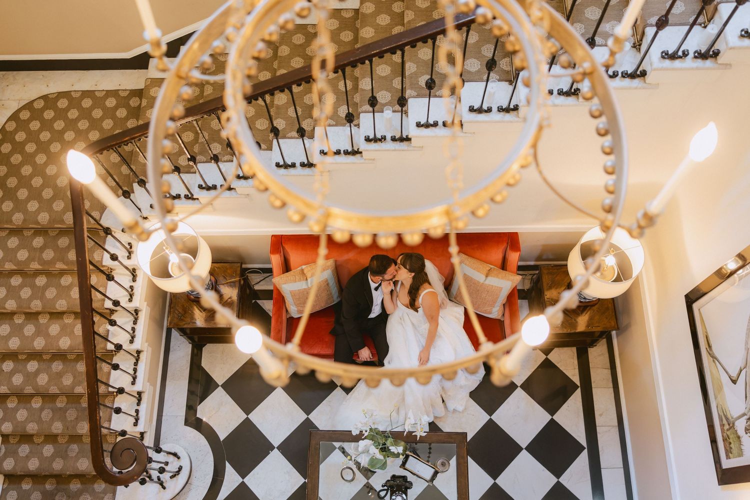 Aerial view of bride and groom kissing on checkered floor beneath elegant chandelier.