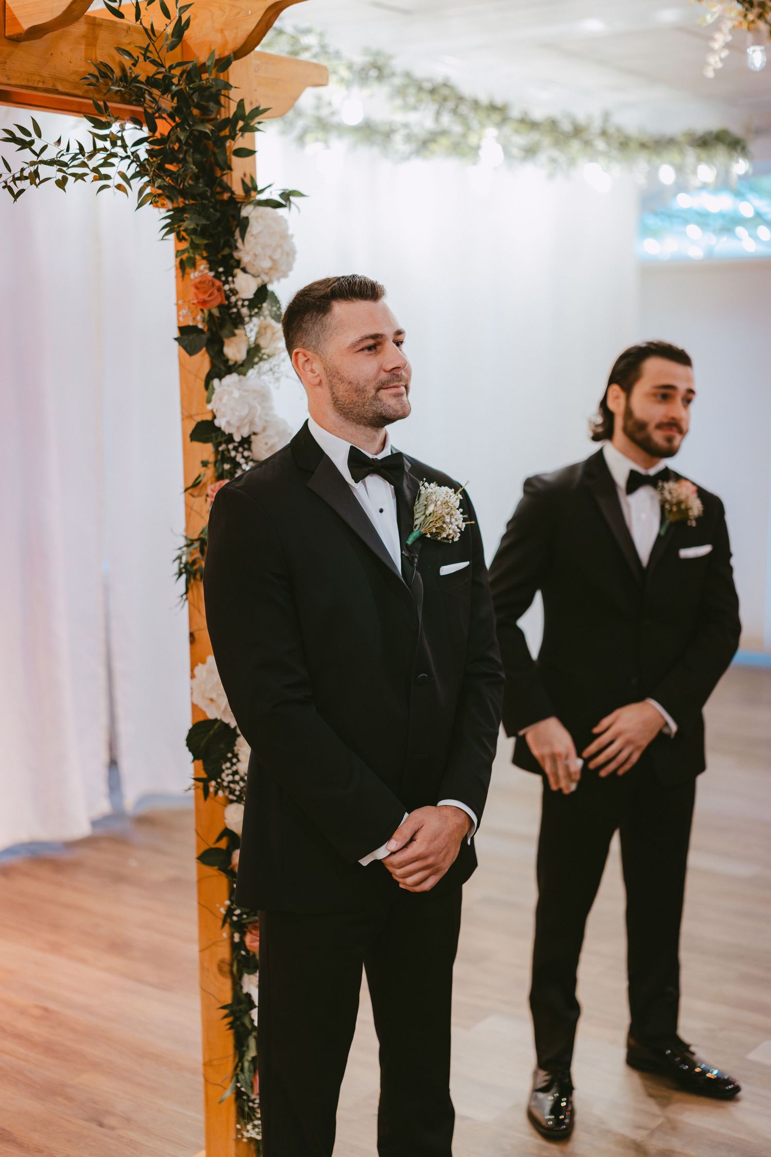 Groom smiles at altar under floral wooden arch with best man beside him at indoor wedding.