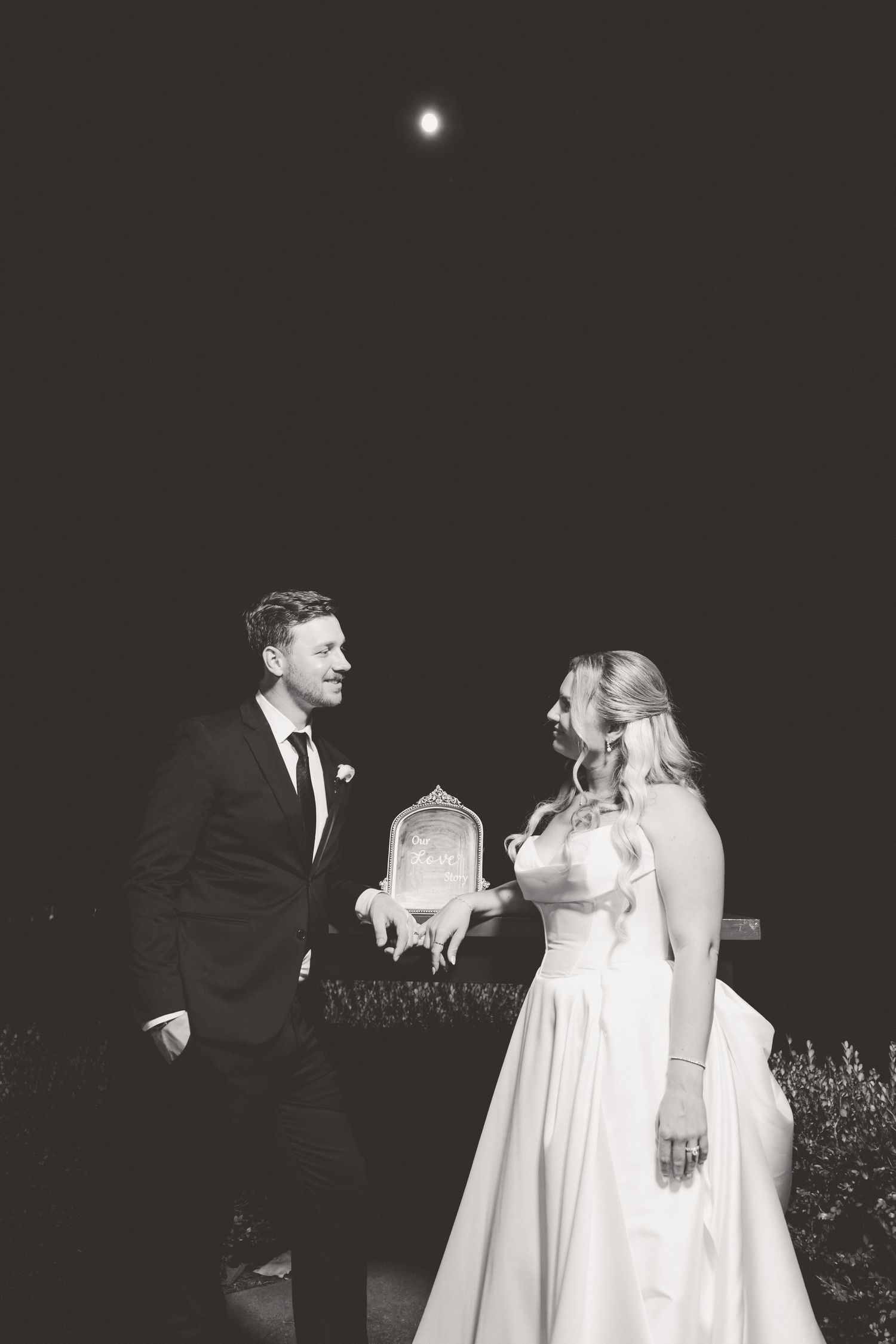Black and white photo of bride and groom sharing a joyful moment outdoors at night under a full moon.