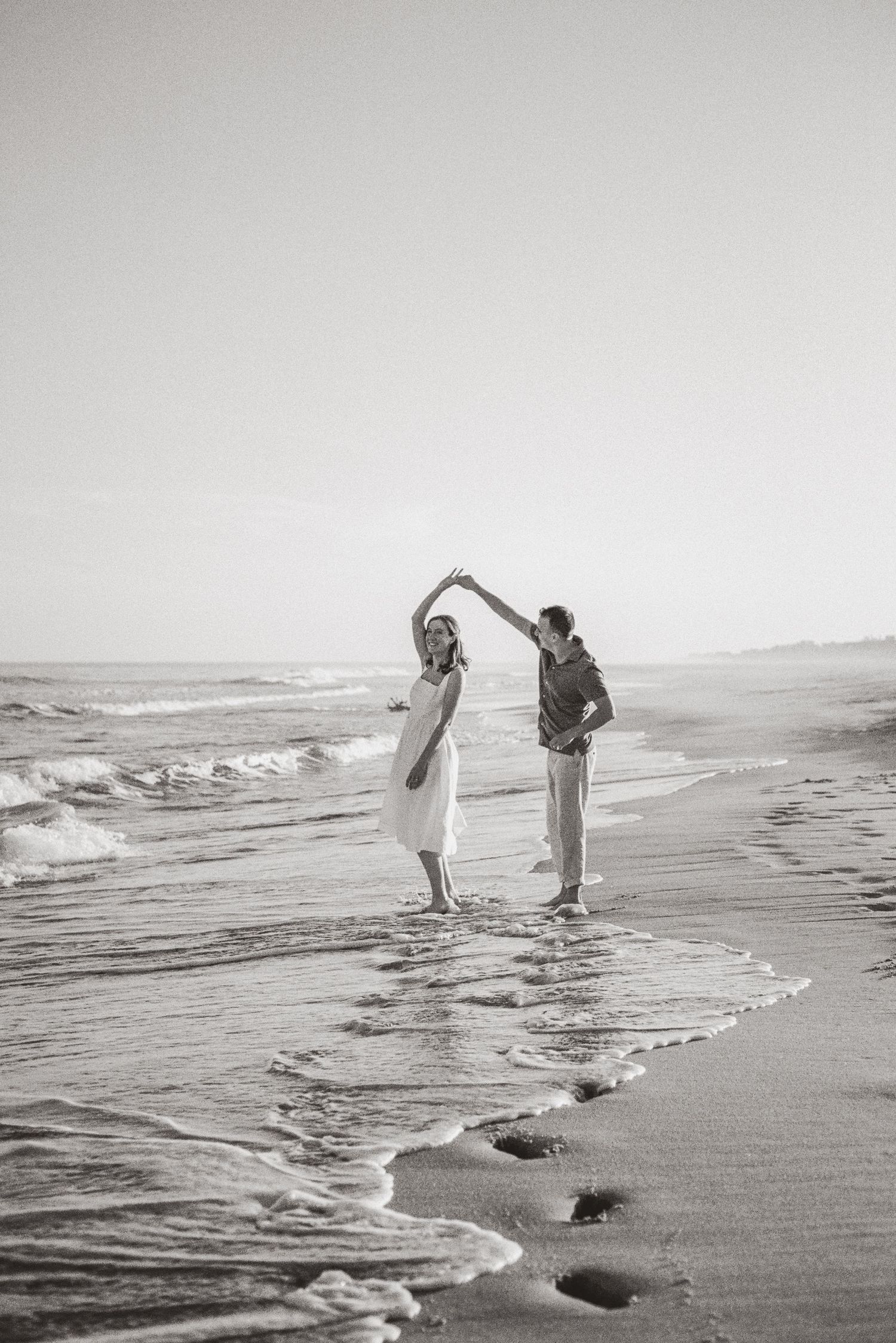 Black and white photo of couple dancing on beach shoreline with ocean waves and footprints.
