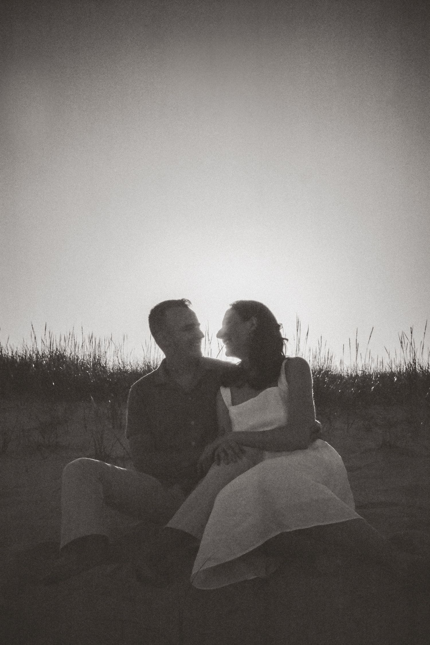Silhouetted couple sitting together on a beach at sunset, smiling at each other in a black and white photo.