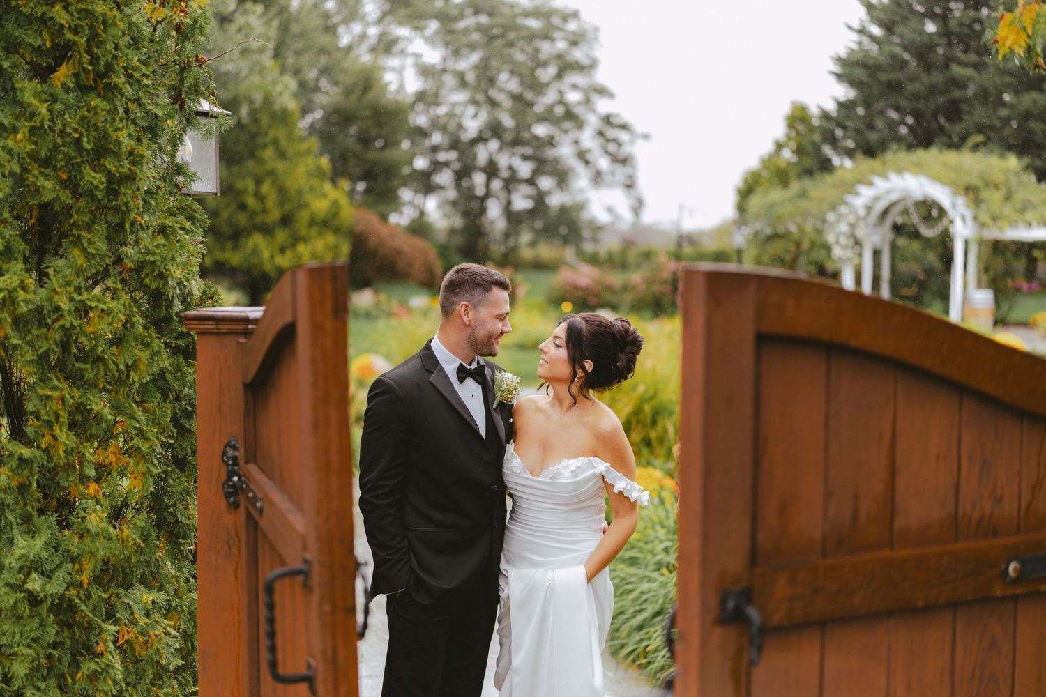 Bride and groom gazing at each other beside rustic wooden garden gates with lush greenery.