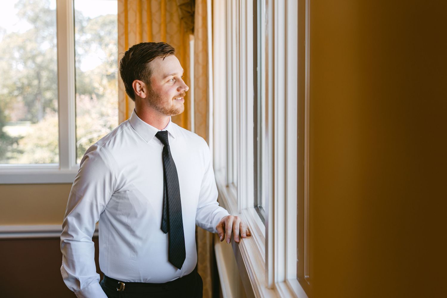 Handsome groom in white shirt and black tie gazing thoughtfully out a sunlit window before wedding.