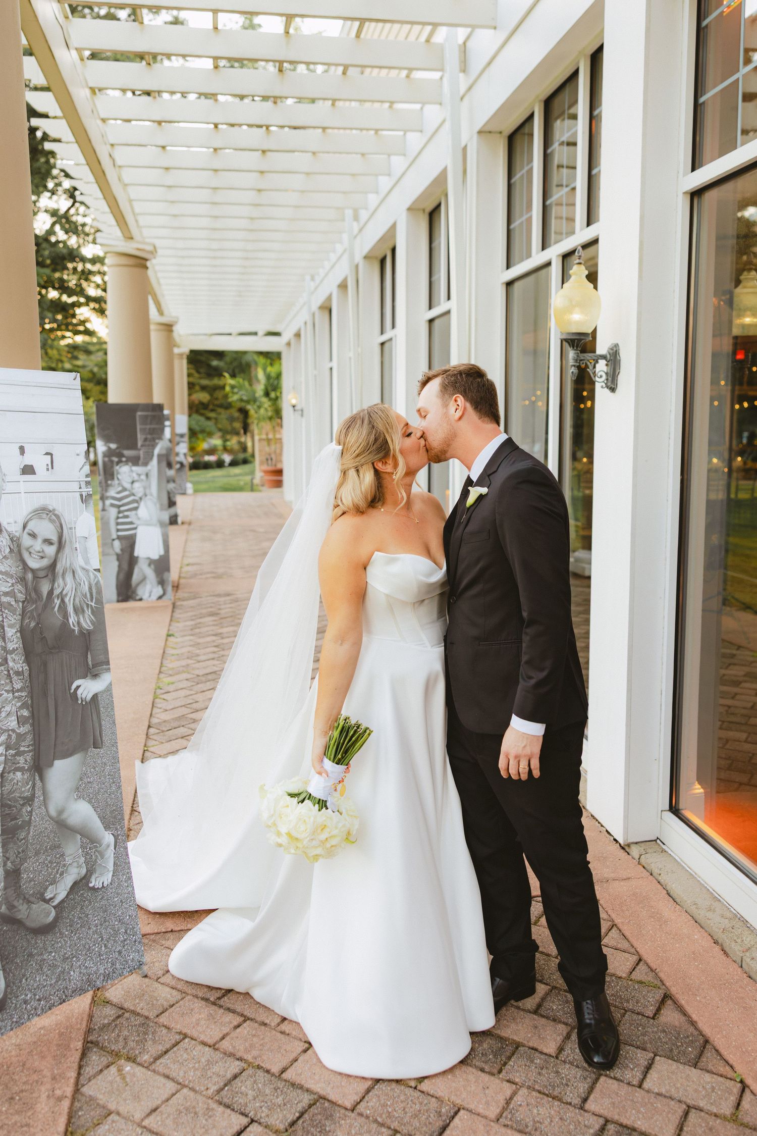 Bride and groom share a kiss under a pergola, bride holding a white floral bouquet in a strapless gown.