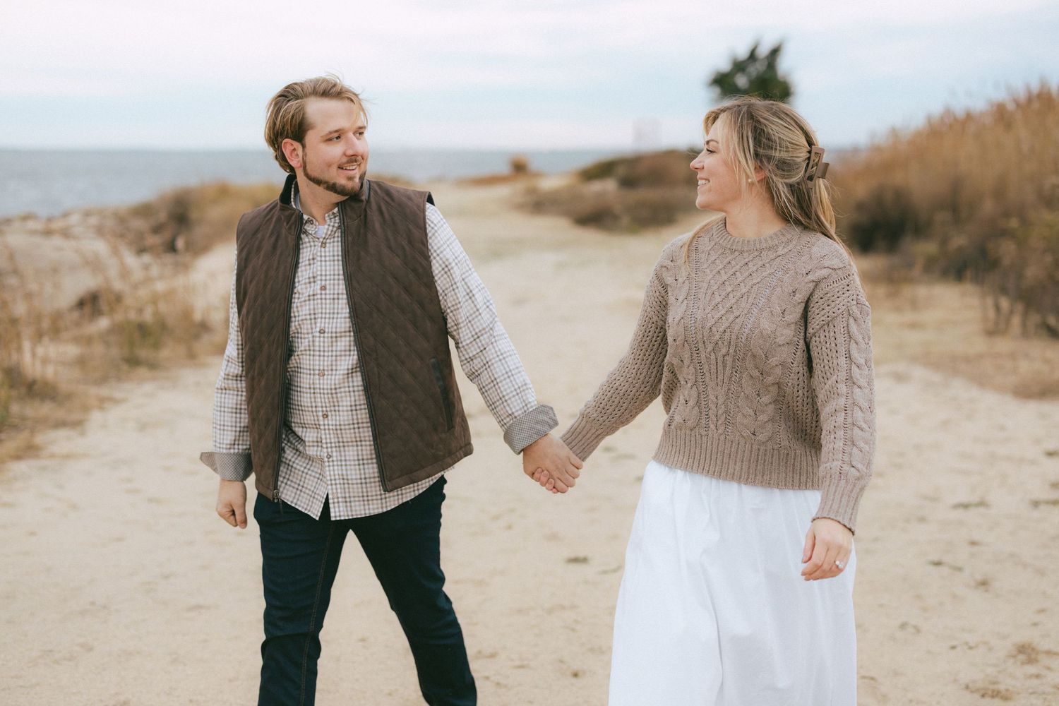 Couple holding hands and smiling at each other on a sandy beach path with tall grass in the background.