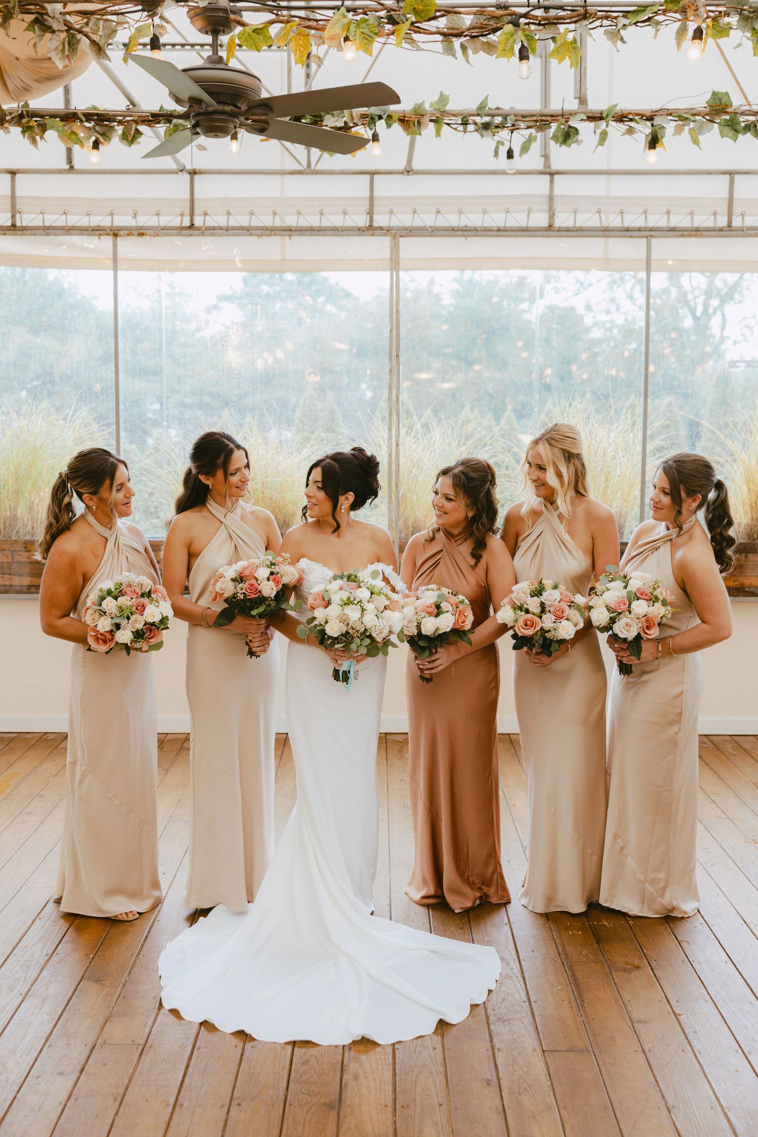 Bride surrounded by five bridesmaids in neutral satin gowns holding matching floral bouquets indoors.