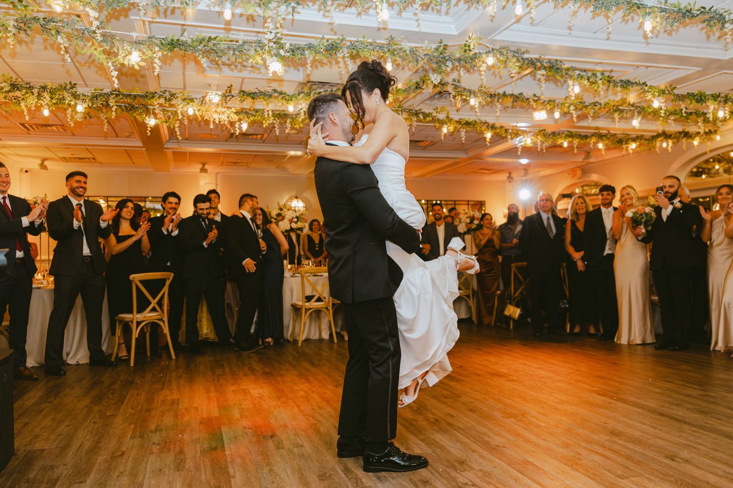 Groom lifts bride during first dance as wedding guests cheer and applaud around them.