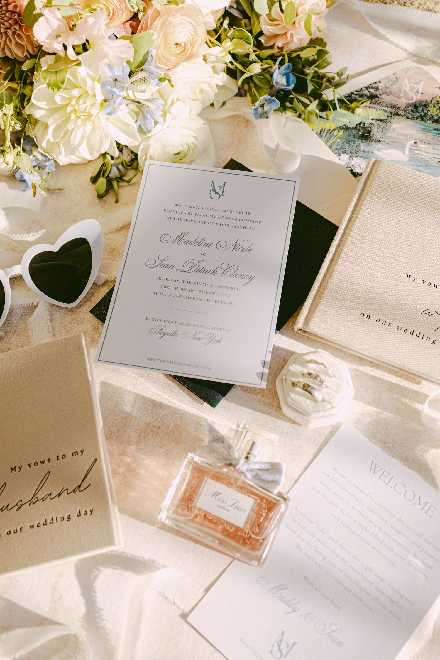 Elegant wedding stationery flat lay with Miss Dior perfume, floral bouquet, and heart sunglasses.