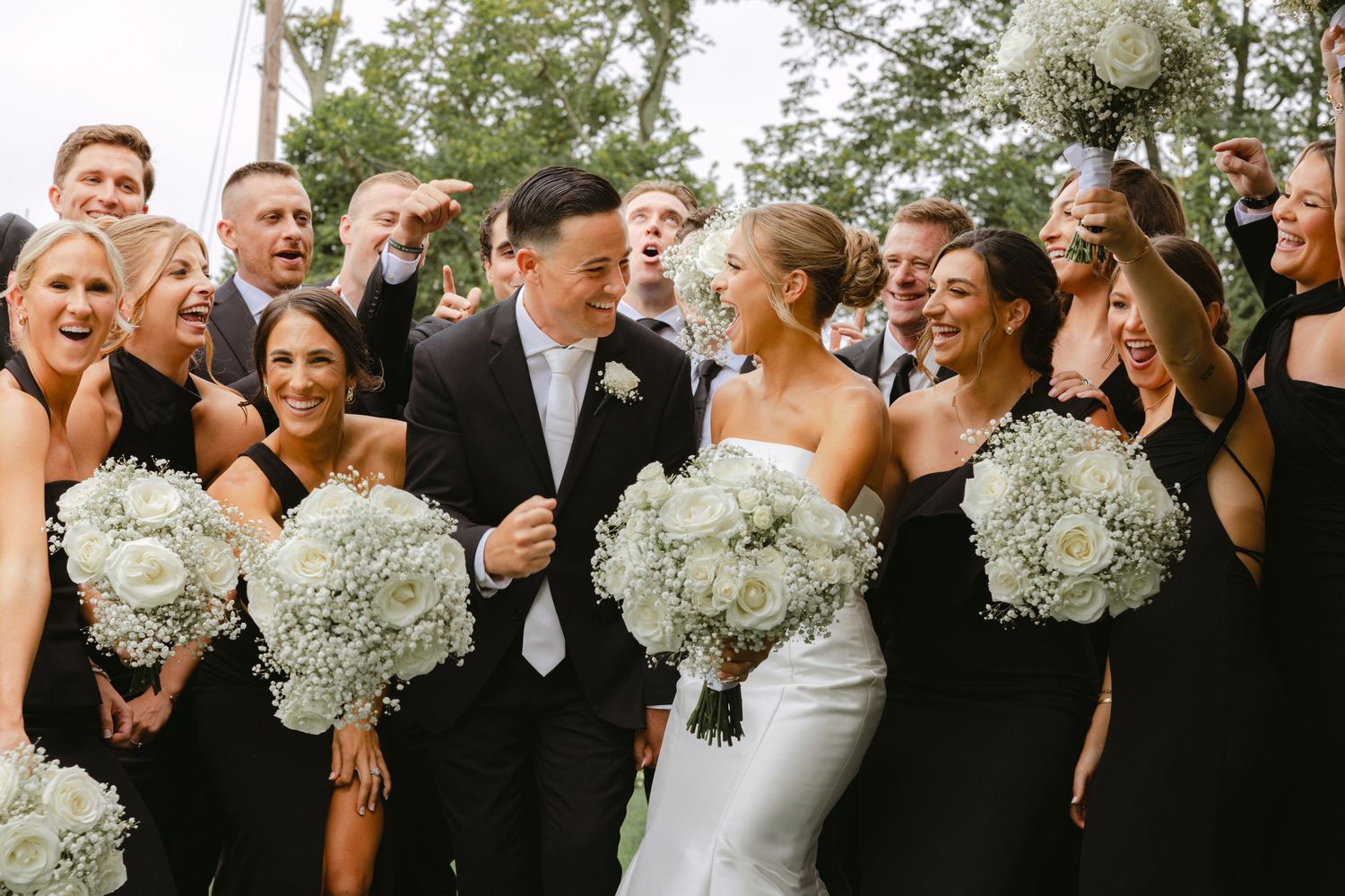 Bride and groom celebrate with wedding party in black attire holding white rose and babys breath bouquets.