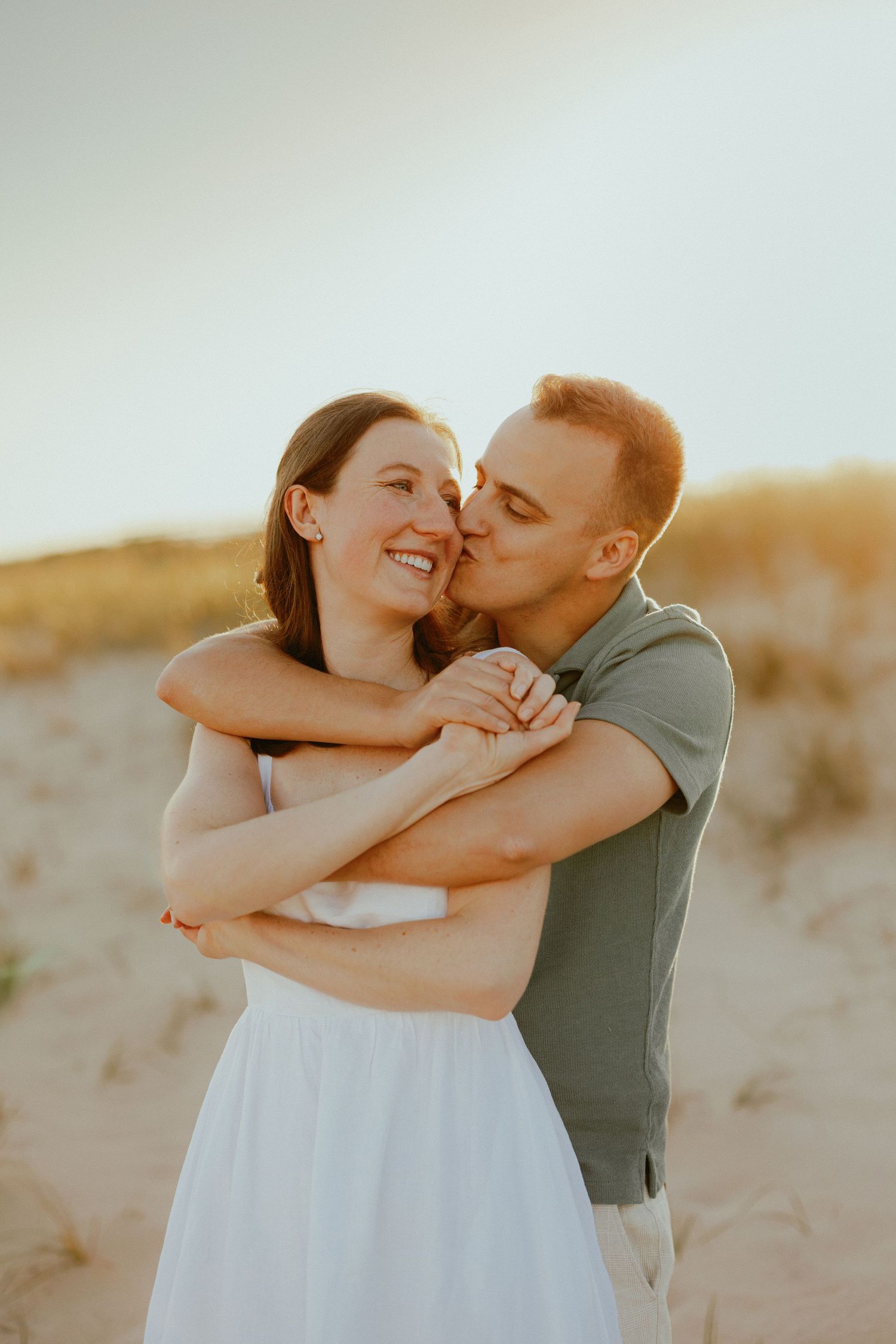 Groom kisses smiling bride on cheek while embracing her on sandy beach dunes at sunset.