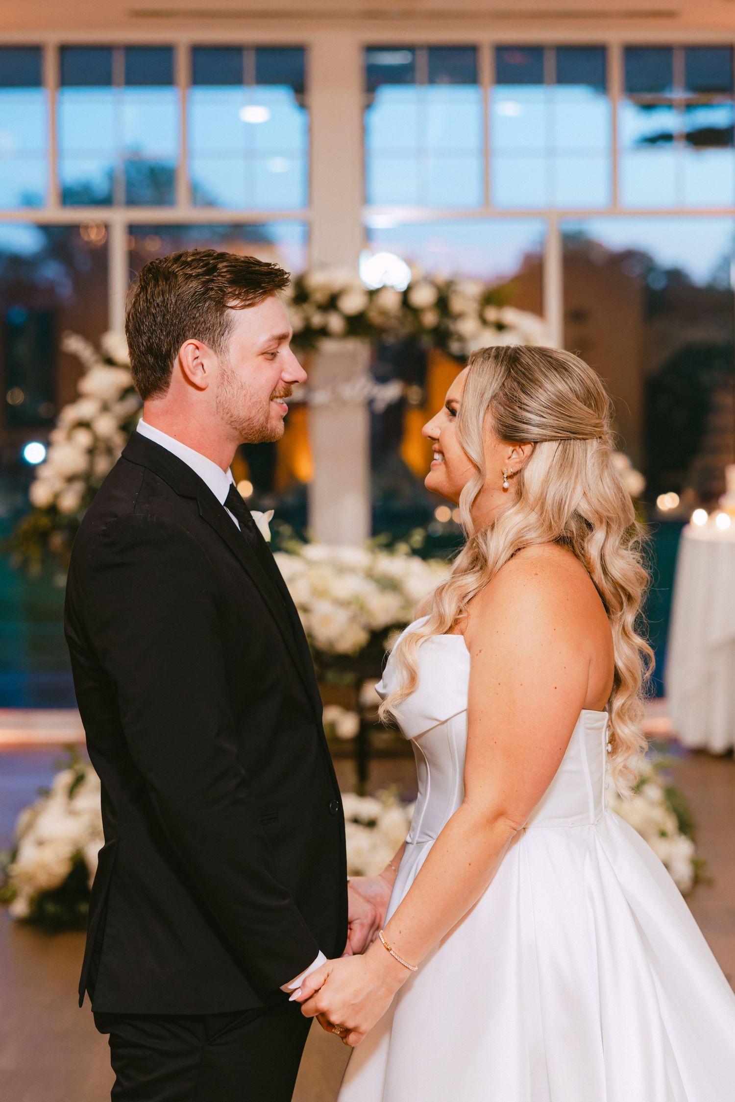 Bride and groom hold hands and gaze at each other in a decorated reception hall with floral arch.