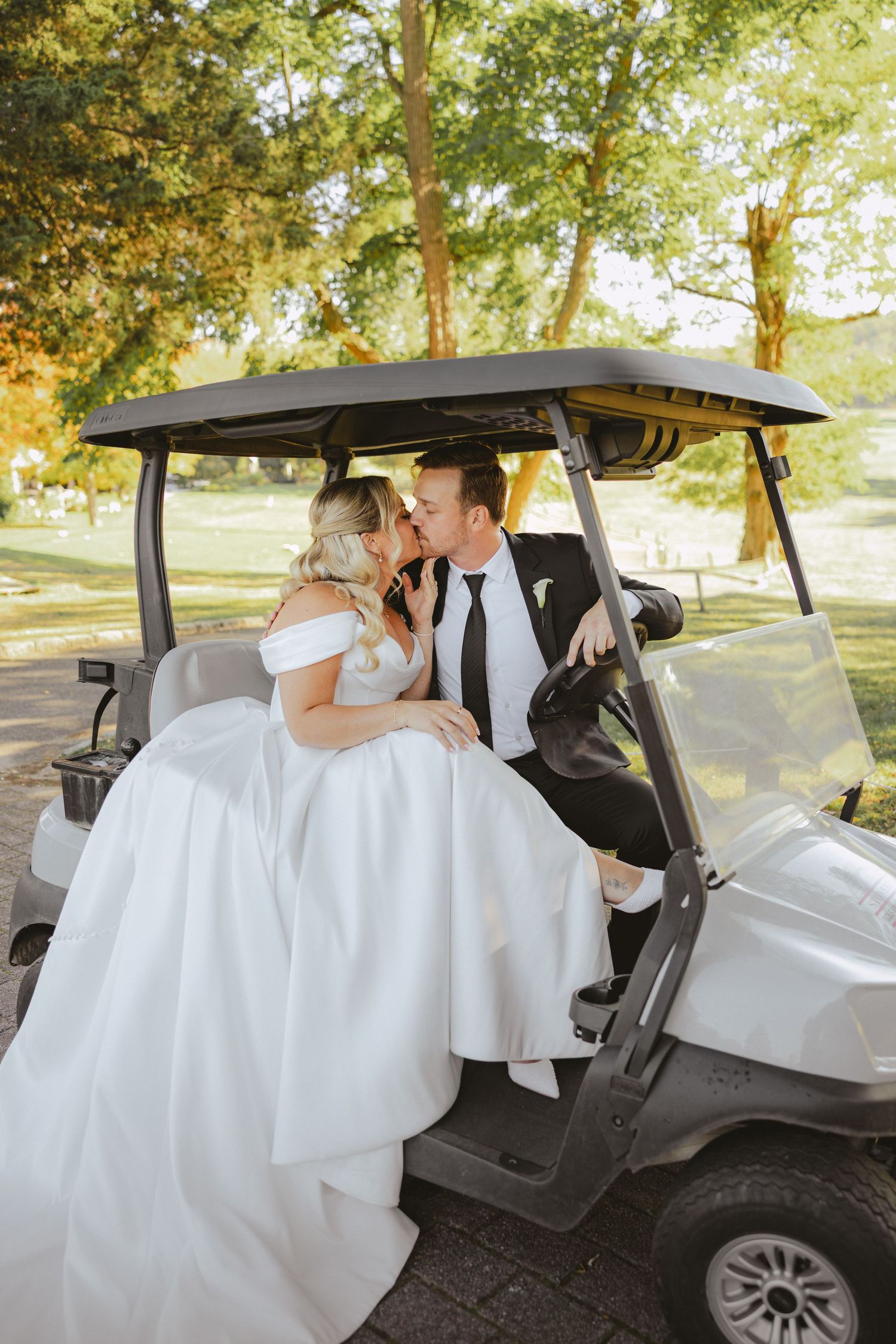 Bride sits on groom lap in a golf cart as they lean in for a kiss in a sunny outdoor setting.