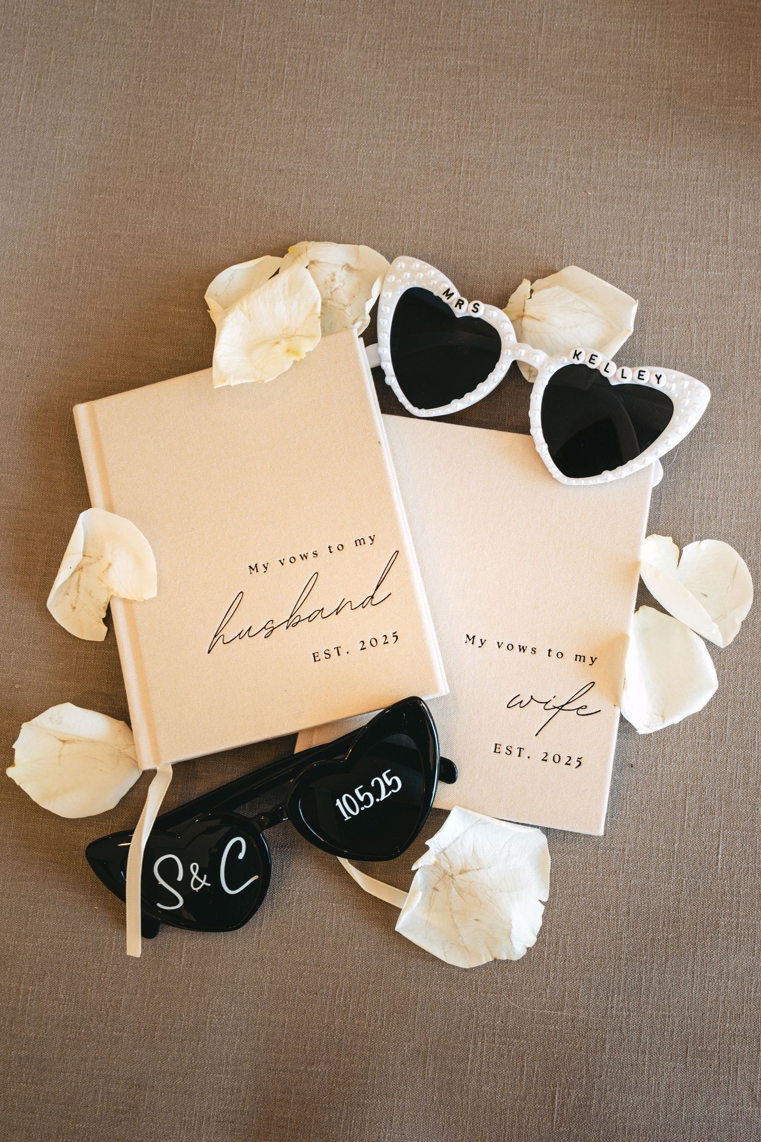 Wedding vow books for husband and wife EST. 2025 with heart sunglasses and white rose petals on neutral background.