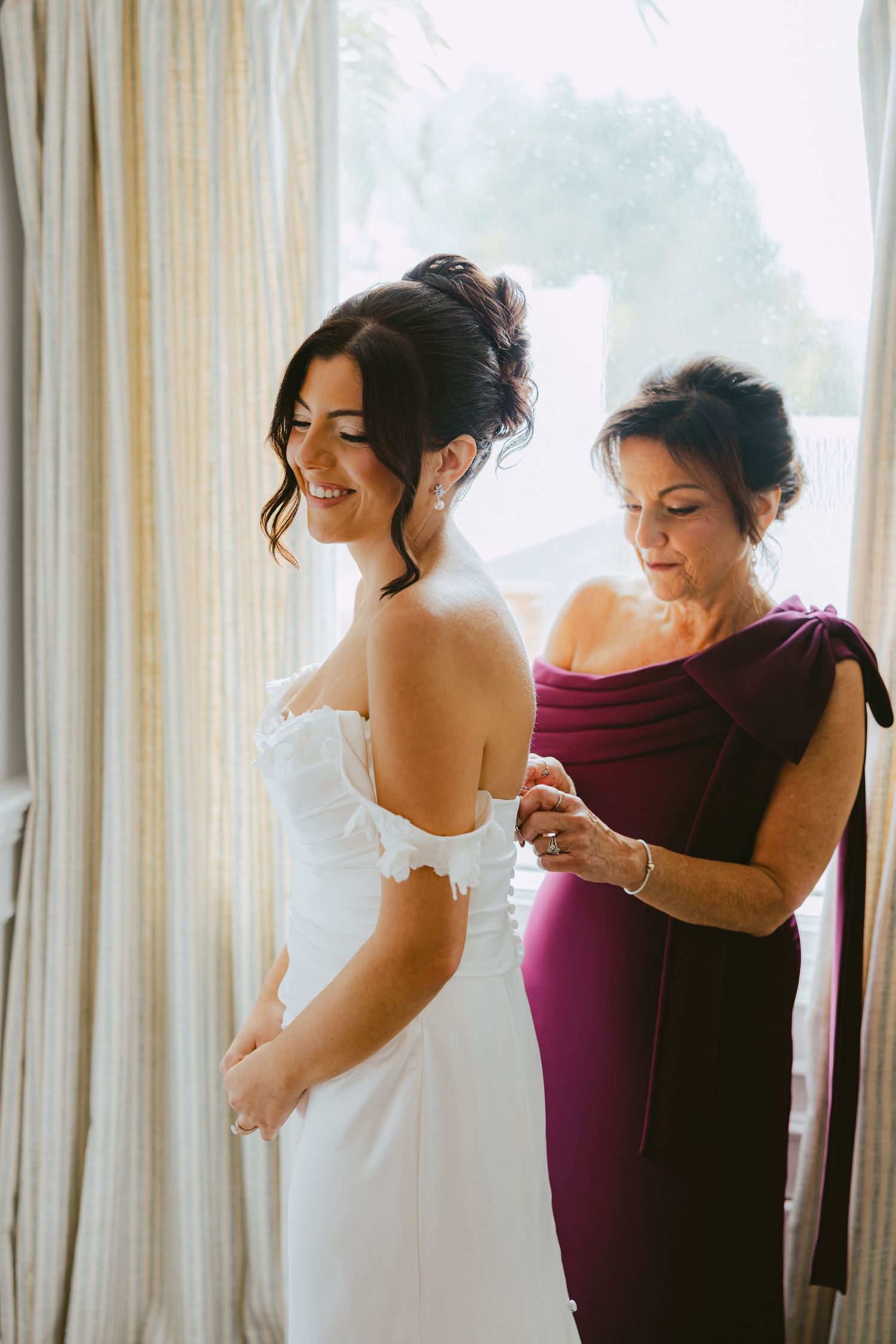 Bride smiling as her mother helps button her white off-shoulder wedding gown near a sunlit window.