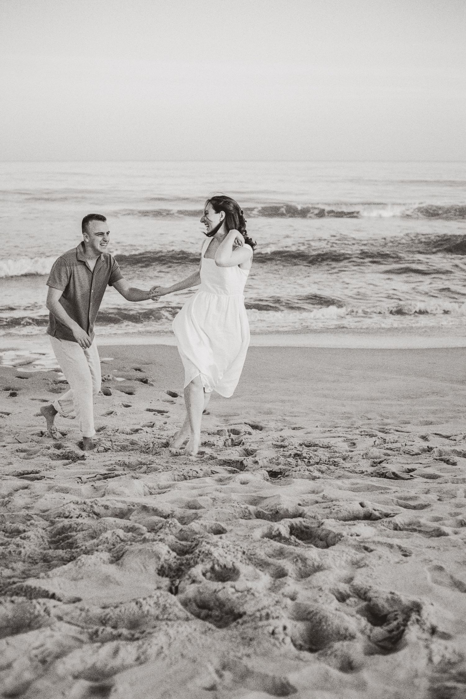 Black and white photo of a couple holding hands and running along a sandy beach with ocean waves.