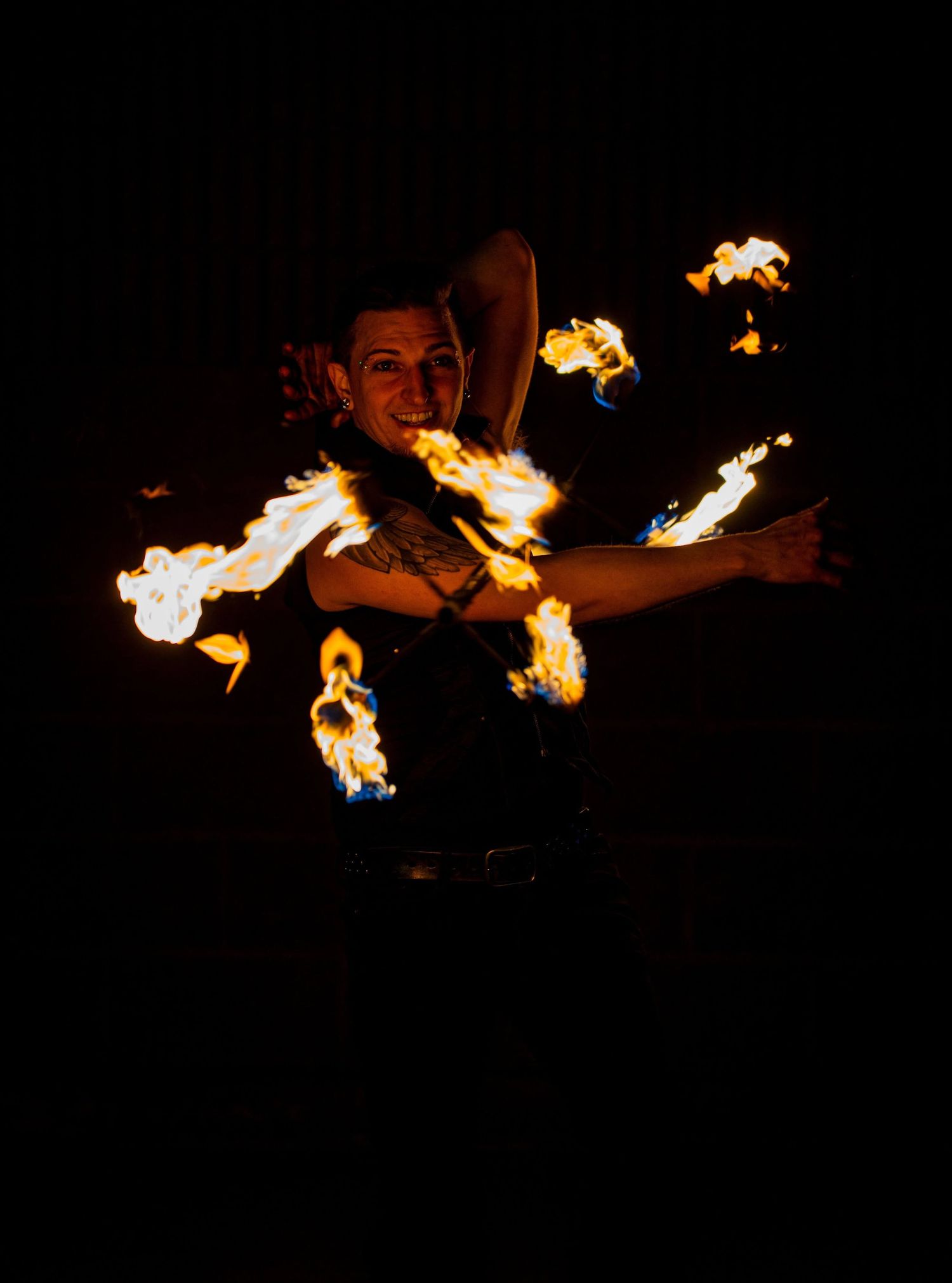A fire dancer performs with multiple flames in the dark, smiling as orange fire illuminates her body.