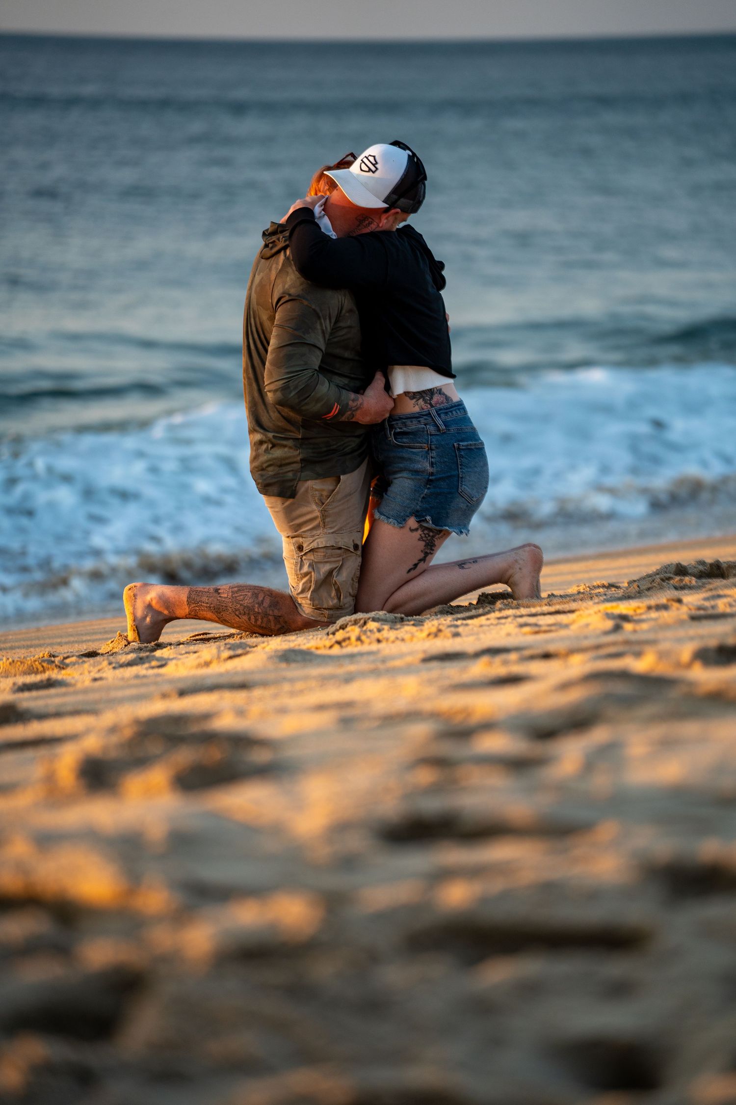 Couple embracing and kissing on a sandy beach at sunset, one person kneeling in the sand as waves roll in behind them.