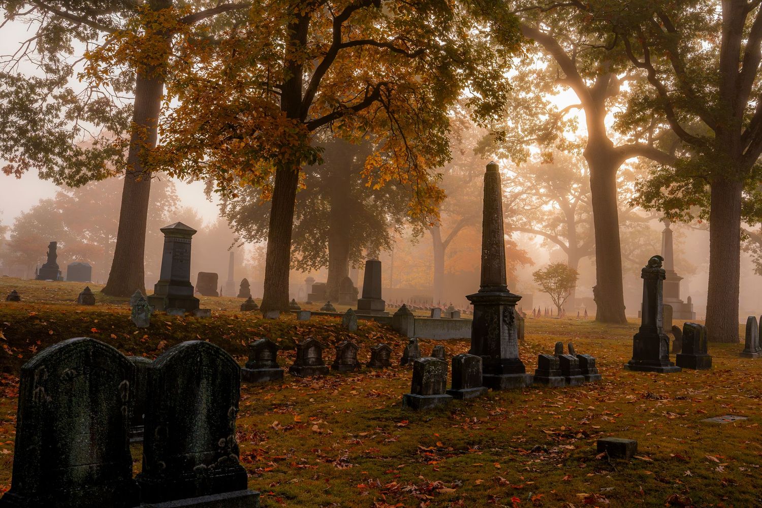 Misty autumn cemetery with historic gravestones, obelisks, and golden foliage trees in soft morning fog.