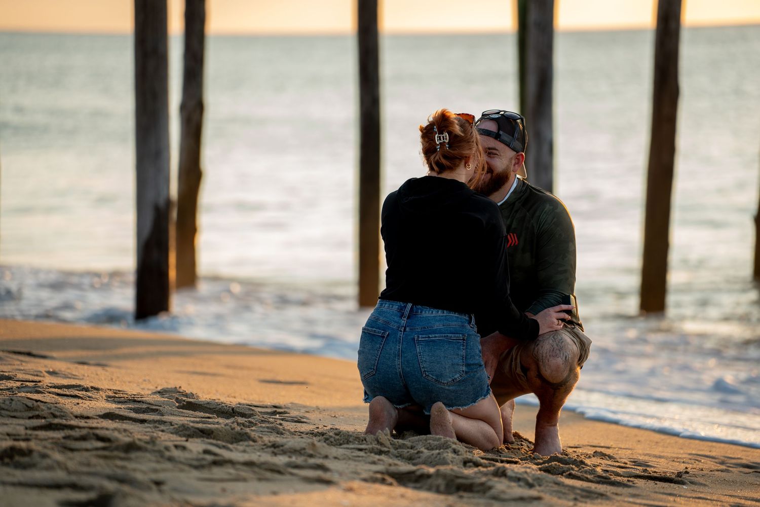 Couple kneeling and embracing on a sandy beach at sunset, with wooden pier pillars and gentle ocean waves in the background.