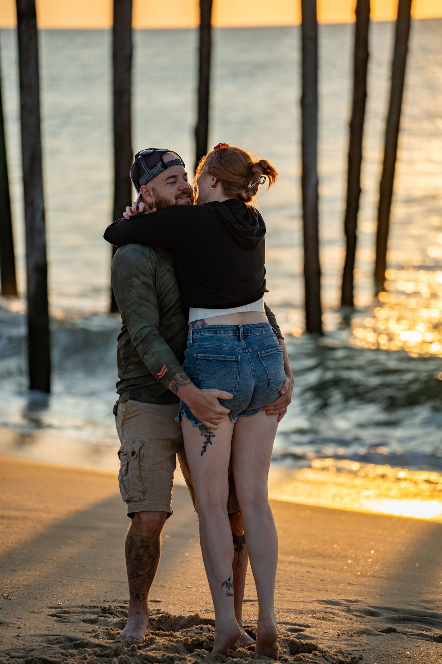 Couple embracing on a sandy beach at sunset, with wooden pier pilings and glowing golden waves in the background.