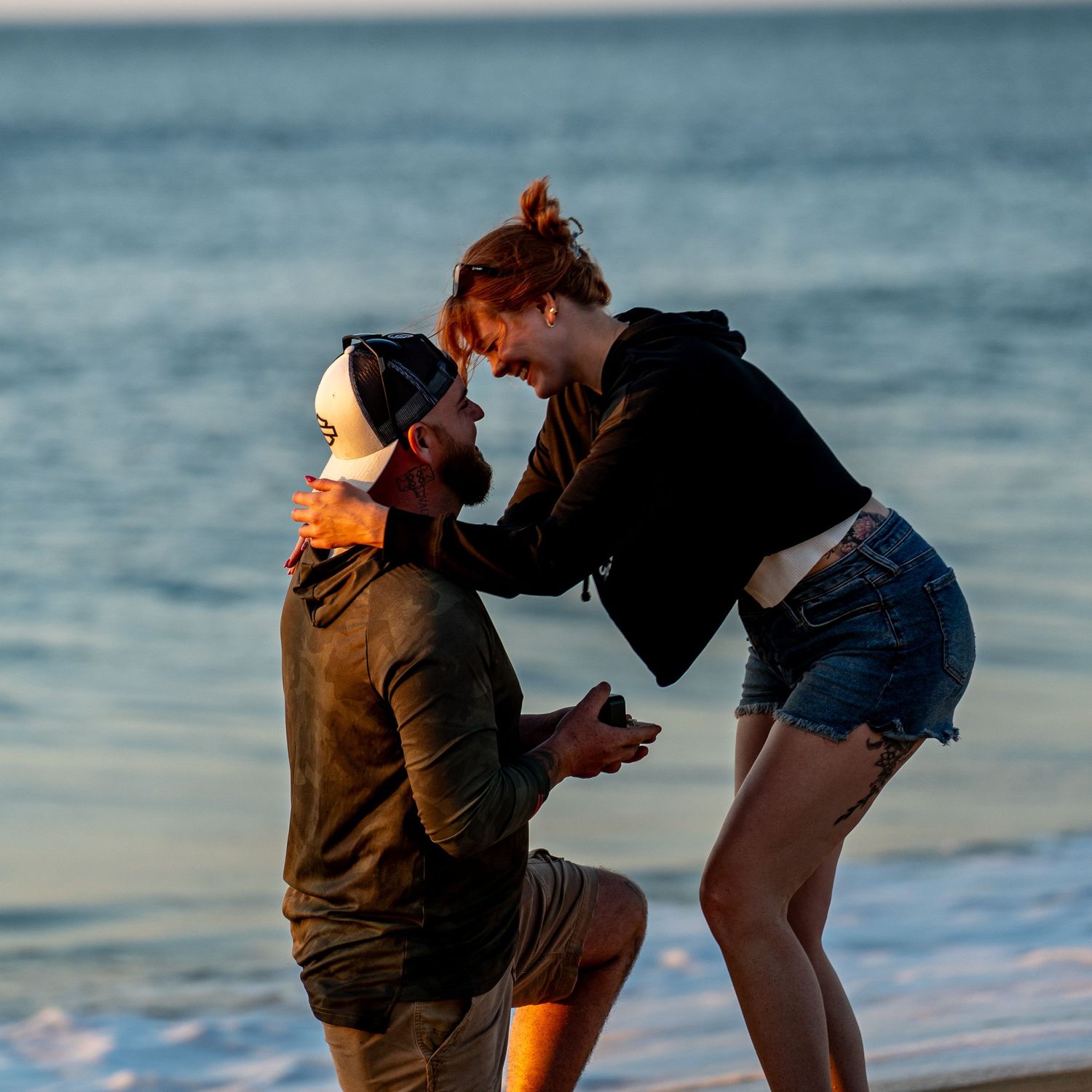 Man in a baseball cap kneels on the beach holding a ring as a smiling woman in denim shorts reaches for him during a sunset proposal.