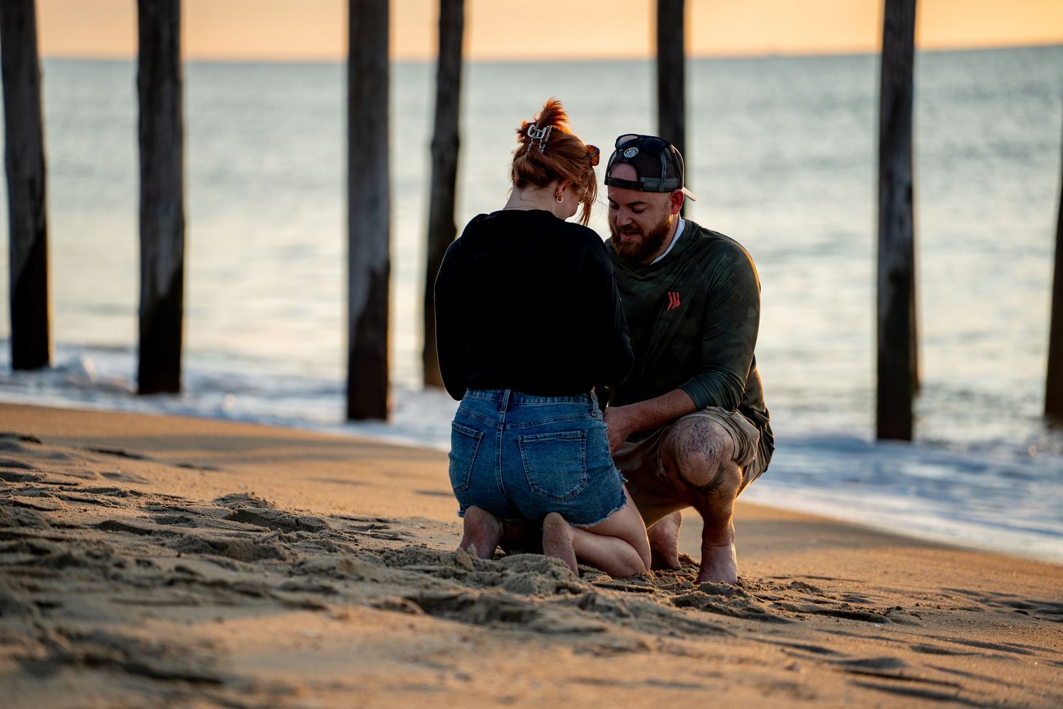 Couple kneeling together on a sandy beach at sunset, with wooden pier pilings and calm ocean waves in the background.