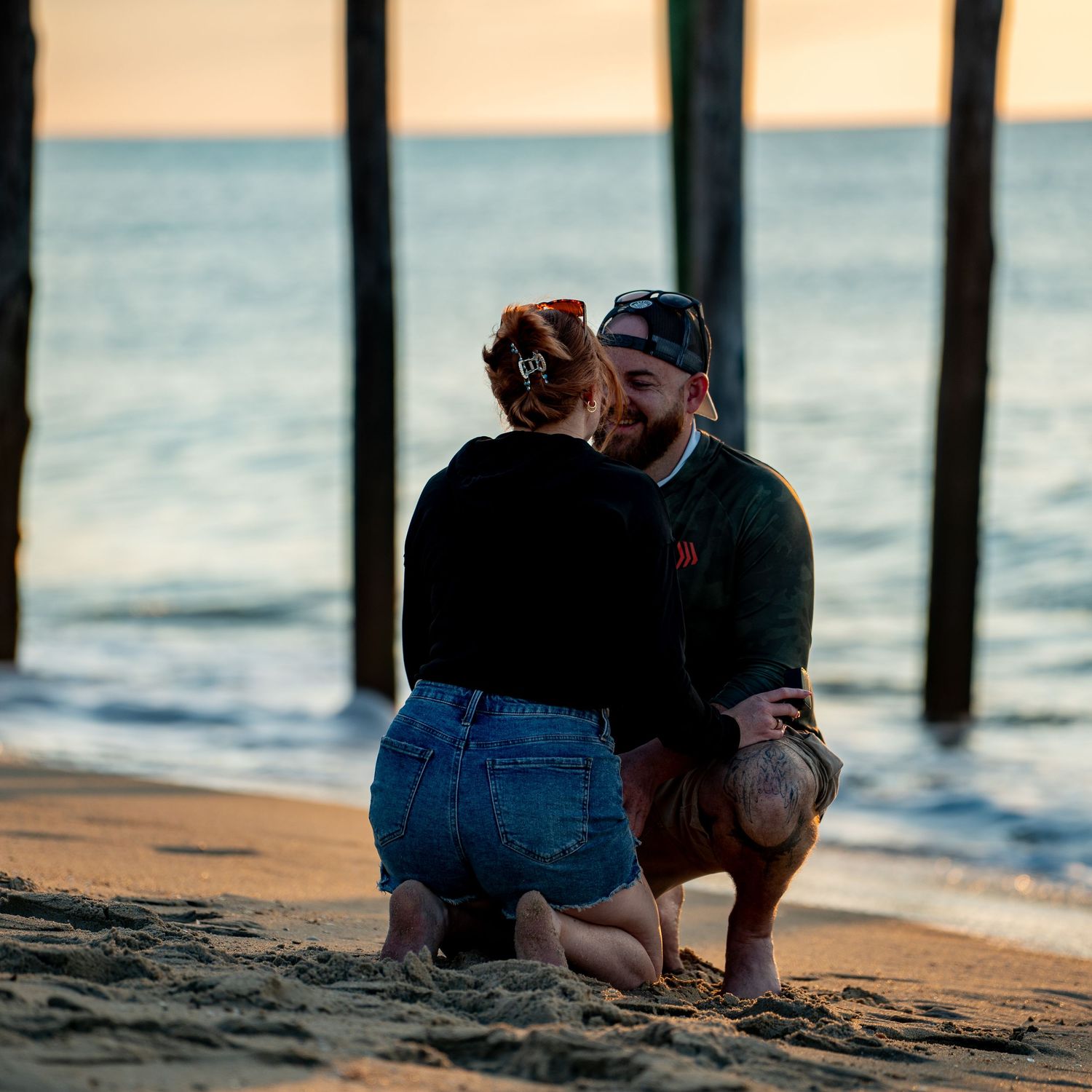 Couple kneeling on a sandy beach beneath wooden pier pilings, sharing an intimate moment as the ocean glows at sunset.