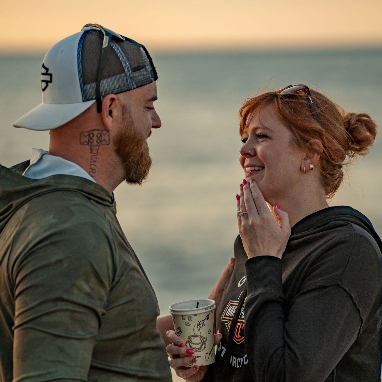 A bearded man in a cap and a smiling red-haired woman holding a coffee cup share a tender moment by the ocean at sunset.